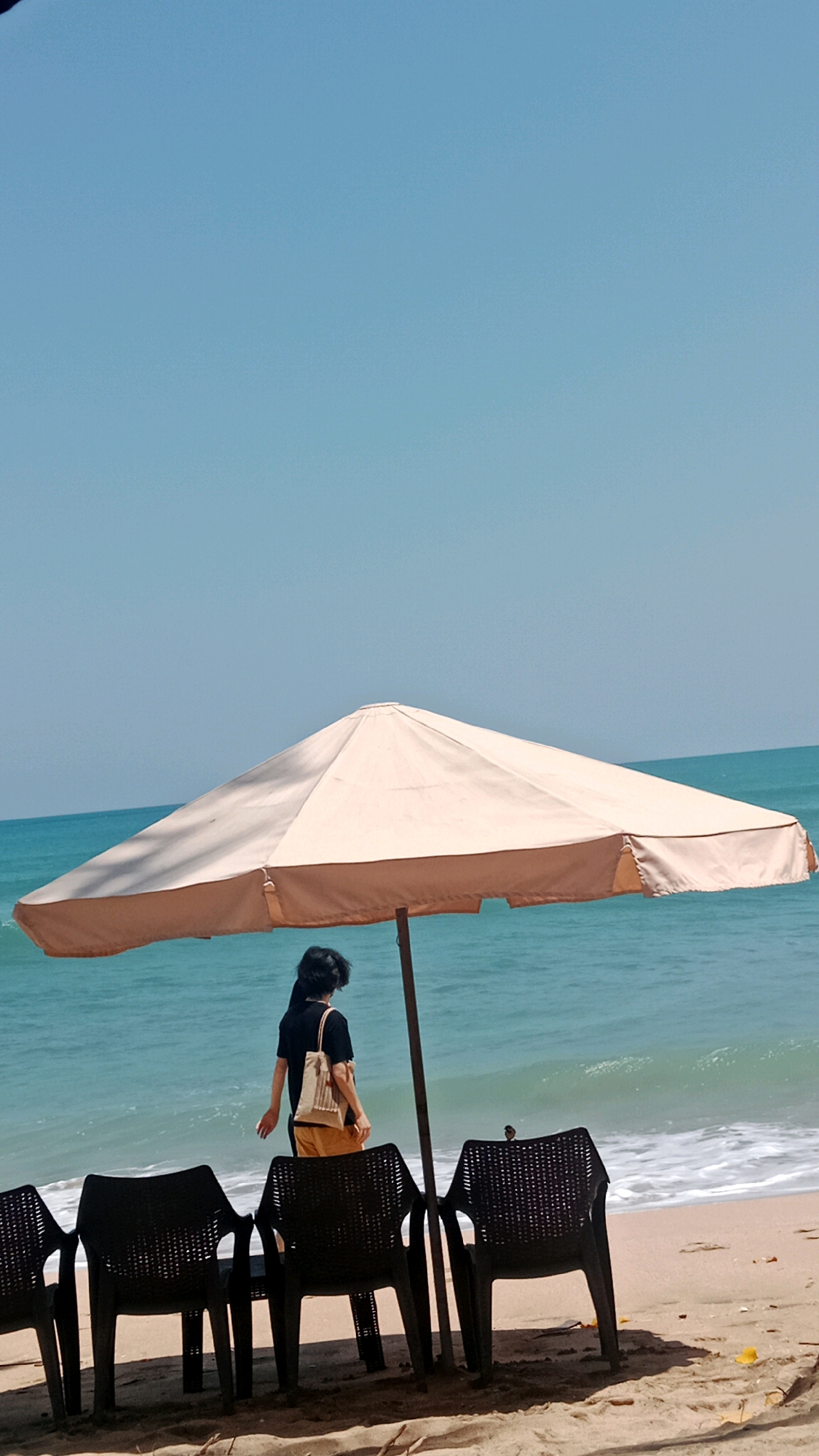 A person stands beneath a large, cream-colored beach umbrella on a sandy shore. The person is facing away from the camera, wearing a dark shirt and light-colored shorts, with a small bag slung over their shoulder. In front of the umbrella are several black plastic chairs. The sea, with gentle waves, is visible behind the umbrella and person. The sky is a clear, pale blue. The location appears to be a beach in Kuta, Indonesia, judging by the tropical setting.