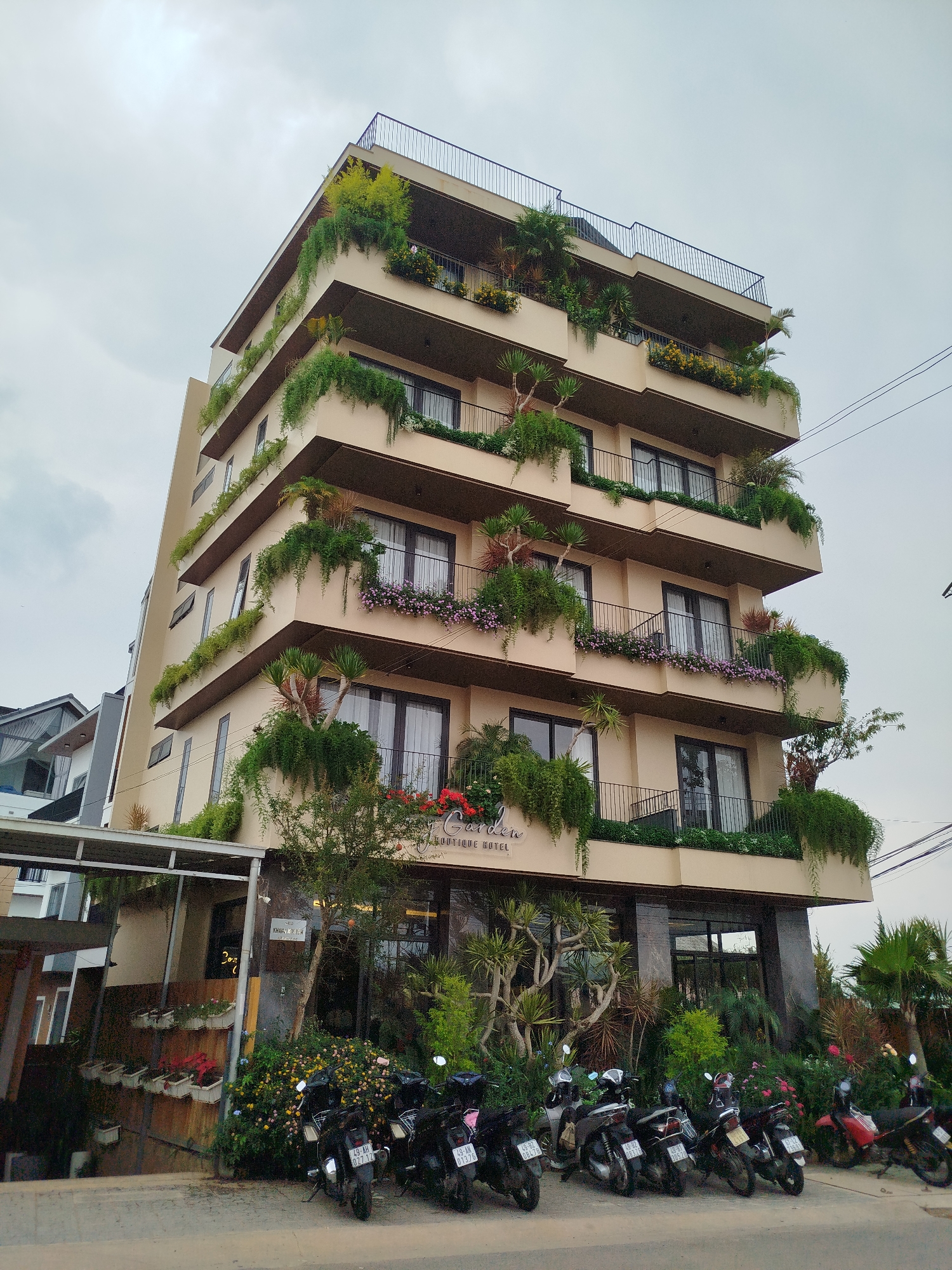The photograph depicts the "J-Garden Boutique Hotel" in Lâm Viên - Đà Lạt, Vietnam. The hotel is a multi-story building with balconies on each level, heavily decorated with greenery and flowers. The building is beige with dark brown window frames and balcony railings. The top floor features a rooftop terrace with a black railing.
At the front of the building, there's dense vegetation including trees and flowers. Several motorcycles are parked in front of the hotel. A neighboring building is partially visible on the left. The sky is overcast.