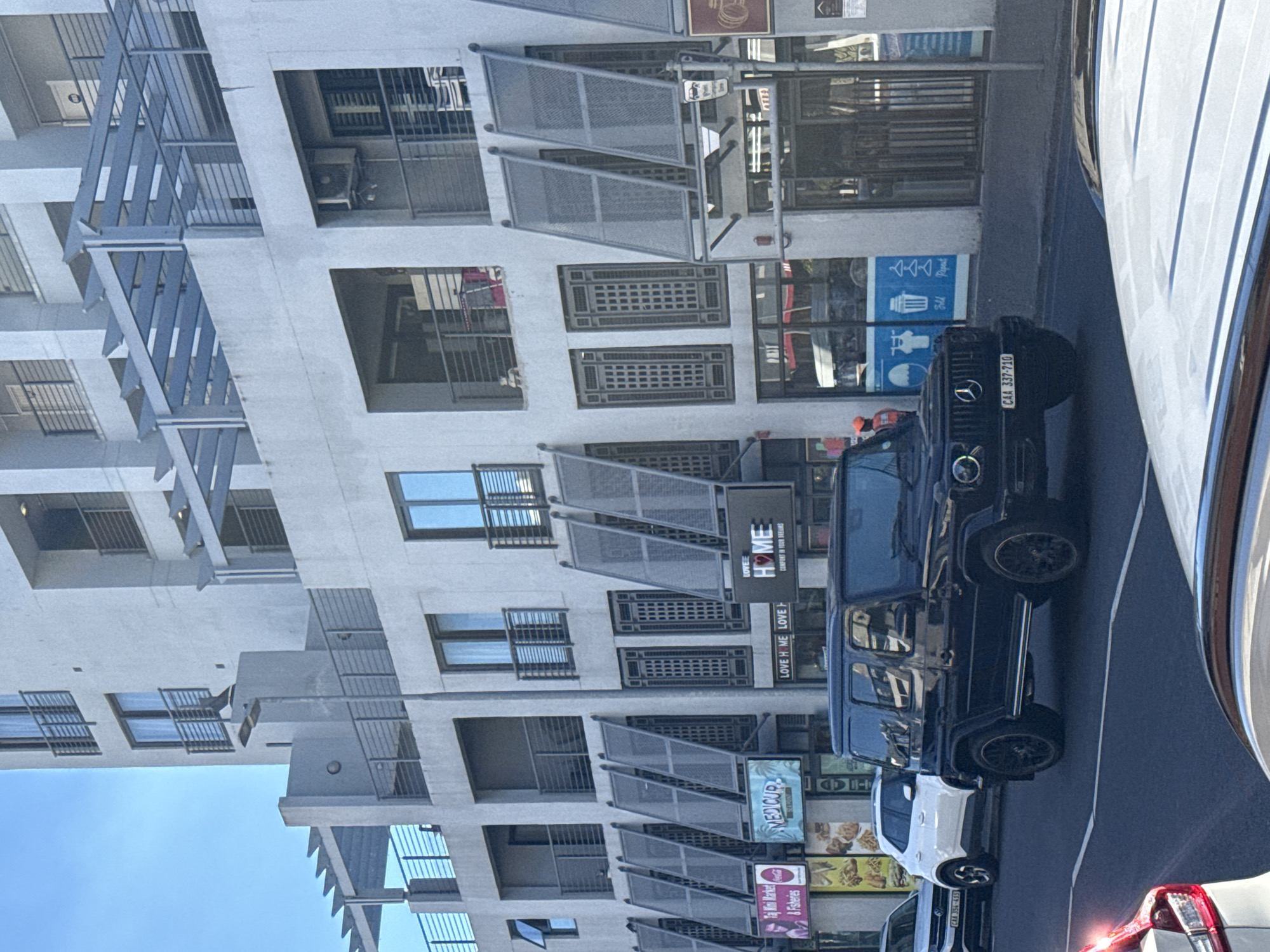 The image shows an urban street scene, likely in Cape Town, South Africa, based on the license plates on the vehicles and the architectural style. A black Mercedes-Benz G-Wagon is parked on the right side of the street, with its license plate "CAA 337-710" visible. Several other cars are parked nearby, including a white sedan. The street is lined with buildings, some appearing to be residential with balconies, and others with retail businesses. Signage for businesses such as "MEDICUP", "LOVEE HOME" and "LOVE H ME LOVE" are present, as well as a sign with the words "Fill Repent". The architecture includes modern elements, such as metal frameworks on the buildings. The photo is taken at an angle, with the buildings appearing tall and the sky visible in the lower left corner. The lighting suggests it is daytime.