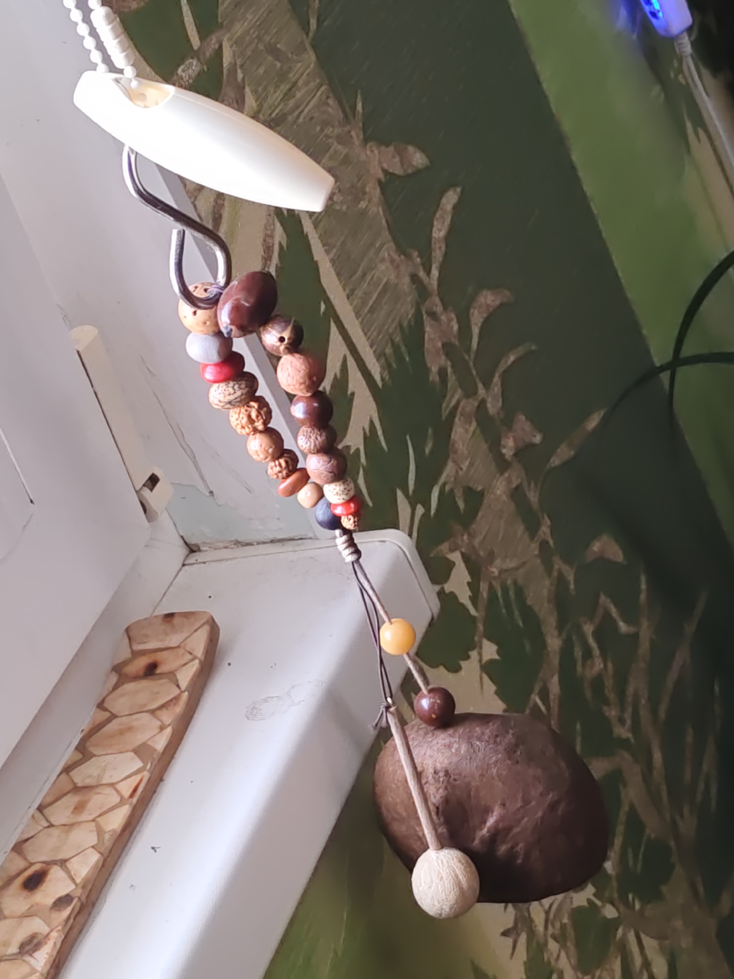 The image depicts a close-up, slightly angled view of a decorative hanging object. This object consists of a white plastic pull for what appears to be a blind or shade, from which hangs a string adorned with various beads. The beads are diverse in color, shape, and material, including natural wood or seed beads, some painted red, and others appearing to be polished stones. The string terminates in a larger, dark brown, irregular-shaped object, possibly a seed pod or a small stone, from which hangs a lighter, spherical bead.
The background is a green wall with a textured pattern that resembles foliage or branches, rendered in shades of green and a lighter, possibly metallic, brown. To the left of the hanging object, a white vertical surface is visible, likely a window frame or wall, with a piece of carved wood attached. This carved wood has a geometric pattern of interlocking shapes.
A small blue light, possibly from a device, is faintly visible in the upper right corner of the image. The overall lighting suggests an indoor setting. The image does not provide any visual information that would allow for the determination of its location in the city of Rostov-on-Don, Russia.