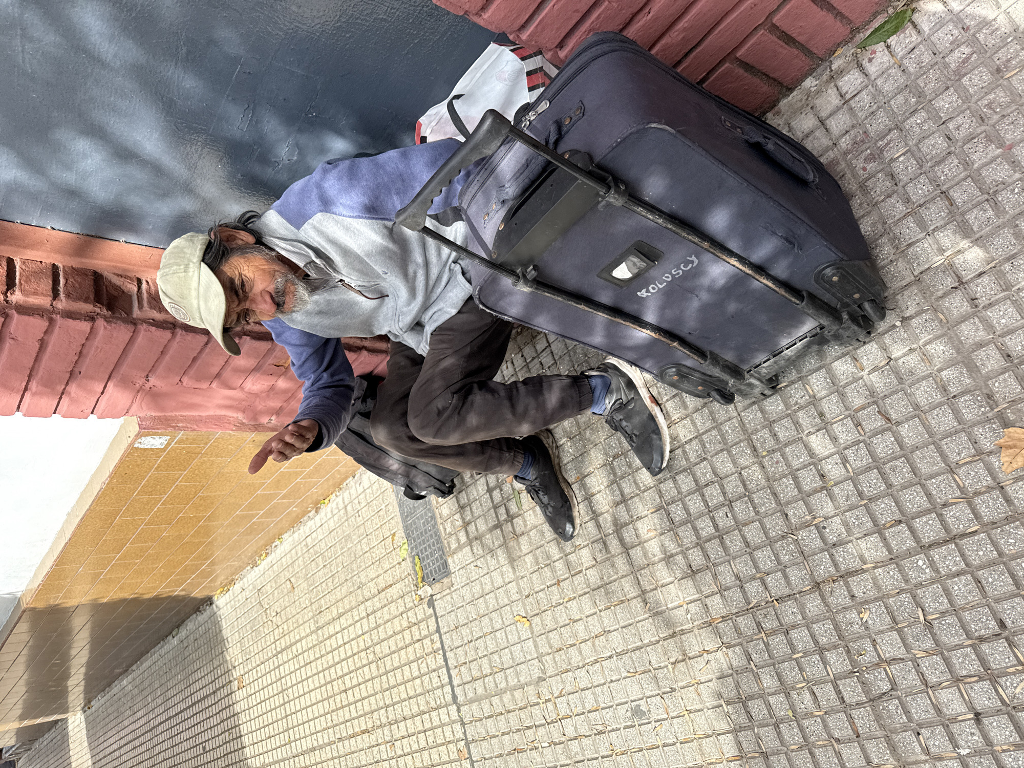 An image, captured at an oblique angle, shows a man resting on a sidewalk in what appears to be Buenos Aires, Argentina. The man, who has a beard and mustache and appears to be elderly, is wearing a light-colored baseball cap, a blue and grey hooded jacket, dark pants, and dark sneakers. He is leaning against a red brick wall, with his legs bent and knees pulled up. A dark blue wheeled suitcase is positioned next to him, its handle extended. The word "KOLOSCY" is written in white letters on the side of the suitcase. A white and red striped shopping bag is also visible behind the suitcase.
The man's right arm is slightly raised with his index finger pointing, although his eyes appear closed or mostly closed, suggesting he is resting or asleep rather than actively gesturing. The setting is an urban sidewalk with distinctive grey square pavers. Shadows cast on the wall and ground indicate bright, daytime conditions. The man's posture and belongings suggest a challenging living situation, possibly homelessness. Behind him, the red brick wall transitions to a darker section above. Further down the street, a building with yellow-brown square tiles is visible. Scattered dried leaves are present on the sidewalk.