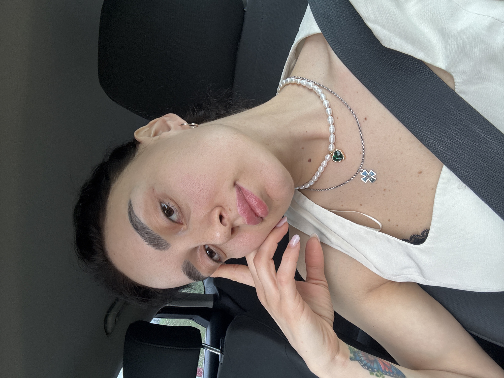 A woman is seated in a car, wearing a white top, a pearl necklace, and a seatbelt. She is holding her face with her hand. A tattoo is visible on her arm. The location is suggested to be Tbilisi, Georgia.