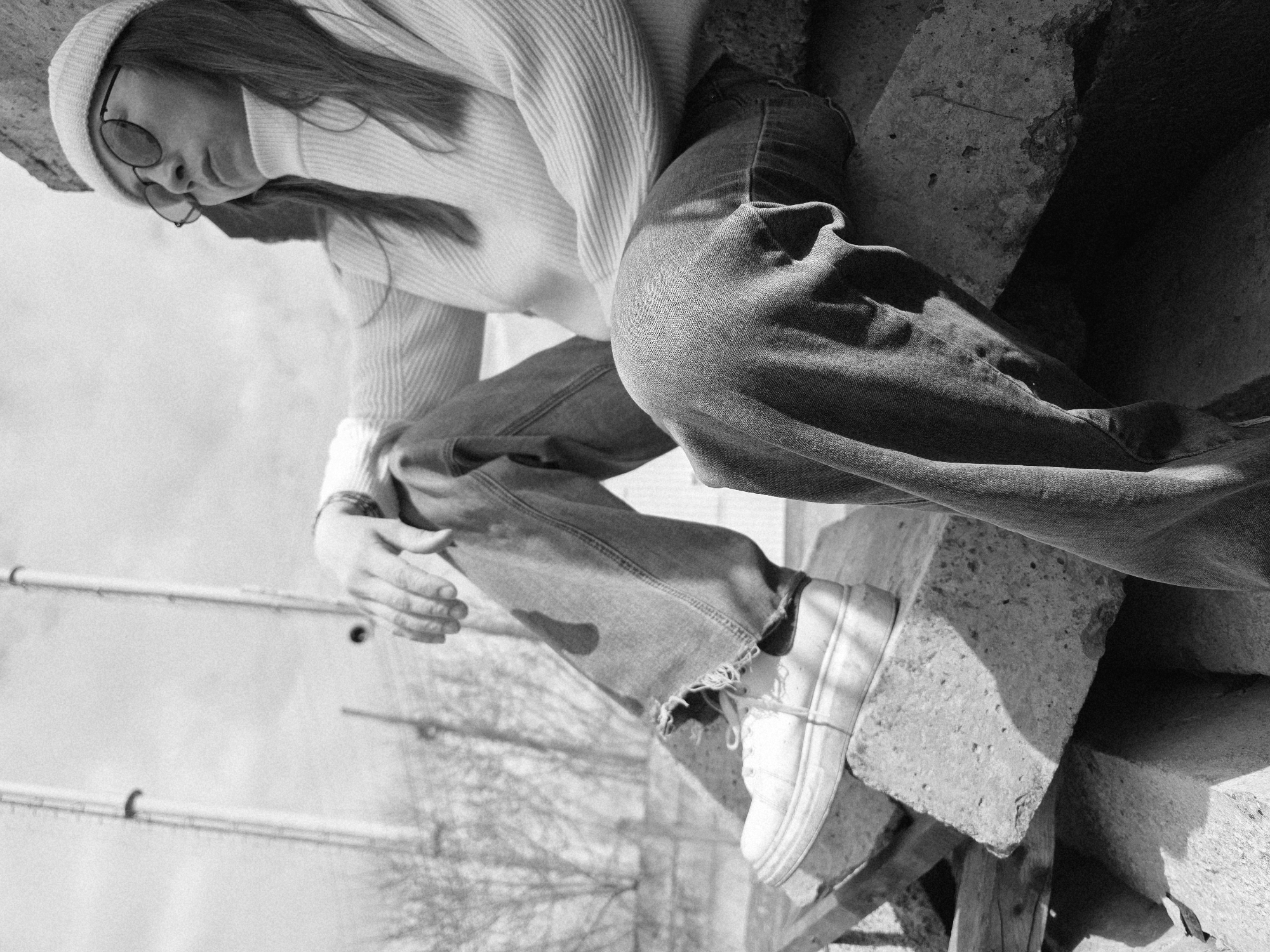 A monochrome photograph depicts a person in a casual attire composed of a ribbed sweater, wide-leg denim pants, and sneakers. The individual is wearing round glasses and a knit cap. They are perched on concrete structures, with their posture suggesting a moment of contemplation or rest. A skeletal tree is visible in the background. The photograph is taken in an outdoor setting that includes architectural elements, possibly in an urban environment.