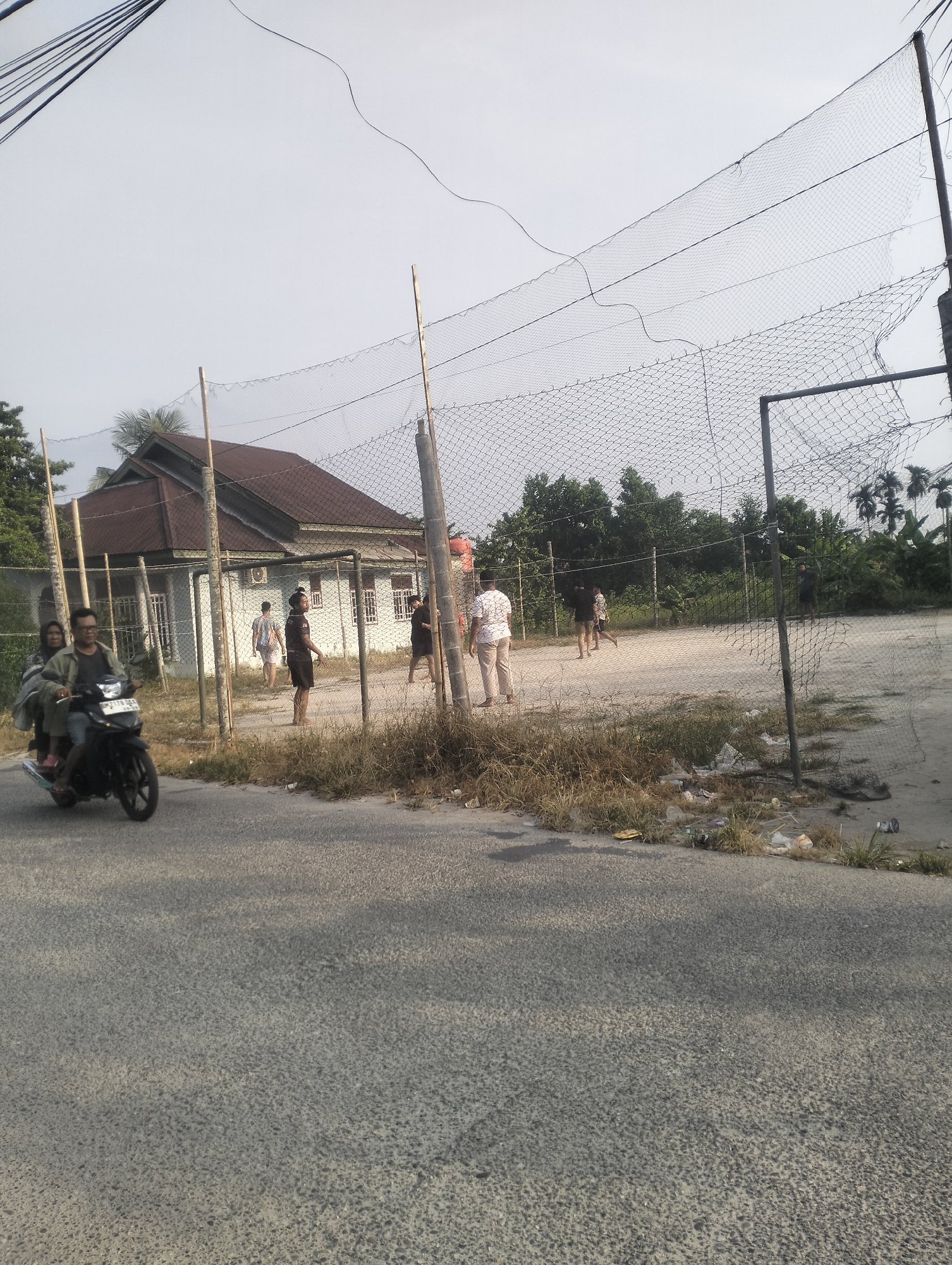 A still image shows a street scene with a motorcycle in the foreground, carrying two people. Behind them, a court area is visible, enclosed by a net and fence, where several individuals are engaged in activities. A house with a brown roof is situated behind the court. The environment appears to be an outdoor setting, possibly in a residential or recreational area within the city of Tambusai Batang Dui, Indonesia. The sky is overcast.