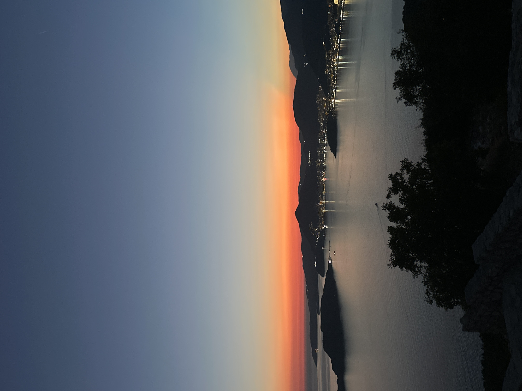The image depicts a twilight scene over a coastal city in Sveti Stefan, Montenegro. The sky transitions from a deep blue at the top to a vibrant orange-red horizon. In the foreground, silhouetted trees and a stone wall create a dark, textured border. The middle ground features a wide expanse of calm water reflecting the faint light. Beyond the water, the cityscape of Sveti Stefan is visible, with numerous lights illuminating the buildings along the coastline and hills. Several small islands or peninsulas are scattered in the water, some with lights. The overall impression is one of tranquility and natural beauty as day transitions into night.
