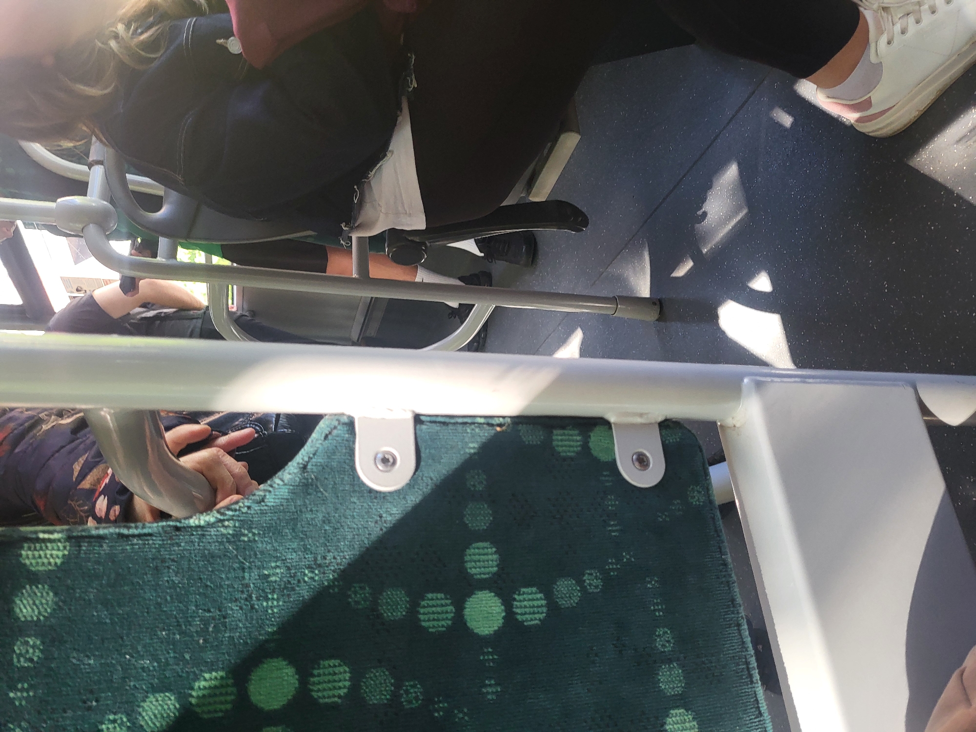 The photograph, taken from a high angle inside what appears to be a public transport vehicle, possibly a bus or tram, depicts parts of two people seated. One person, likely female, wears black leggings, a dark top or jacket and white sneakers, and occupies a seat in the upper right portion of the frame. Another person, wearing a patterned, dark-colored top, sits on a green seat with a circle pattern, positioned in the bottom left of the image. Metallic seat frames and poles are visible, obscuring part of the view. The environment is utilitarian, suggestive of a public space. The city of Sofia, Bulgaria, is not explicitly identifiable within the scene.
