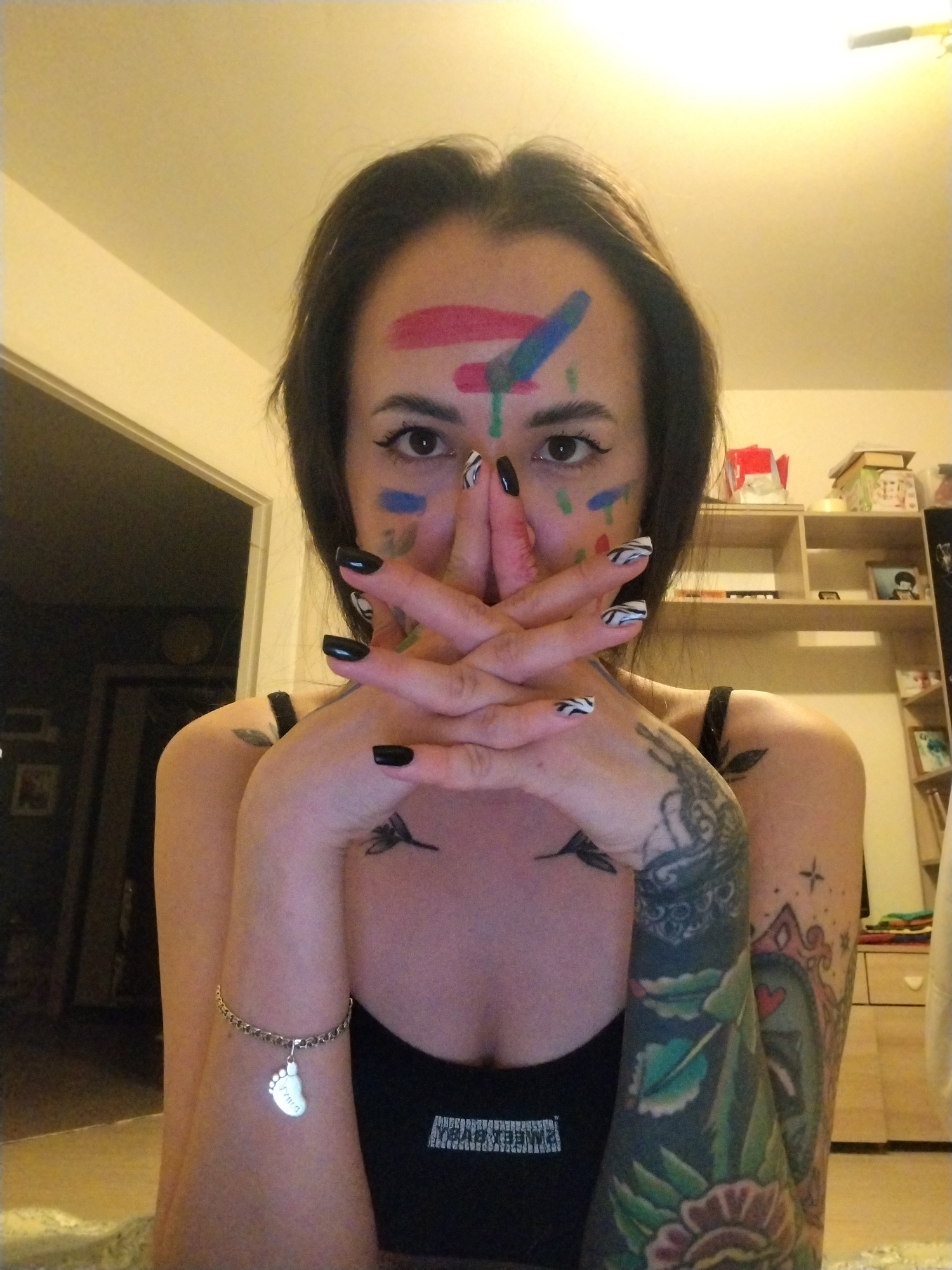 A medium shot depicts a woman indoors. She is positioned in front of a doorway and shelving. She has dark hair, dark eye makeup and paint on her face. Her nails are painted black and with zebra stripes. She is wearing a black tank top. Her left arm is heavily tattooed and she is wearing a bracelet with a foot-shaped charm. Her hands are raised to cover most of her face.