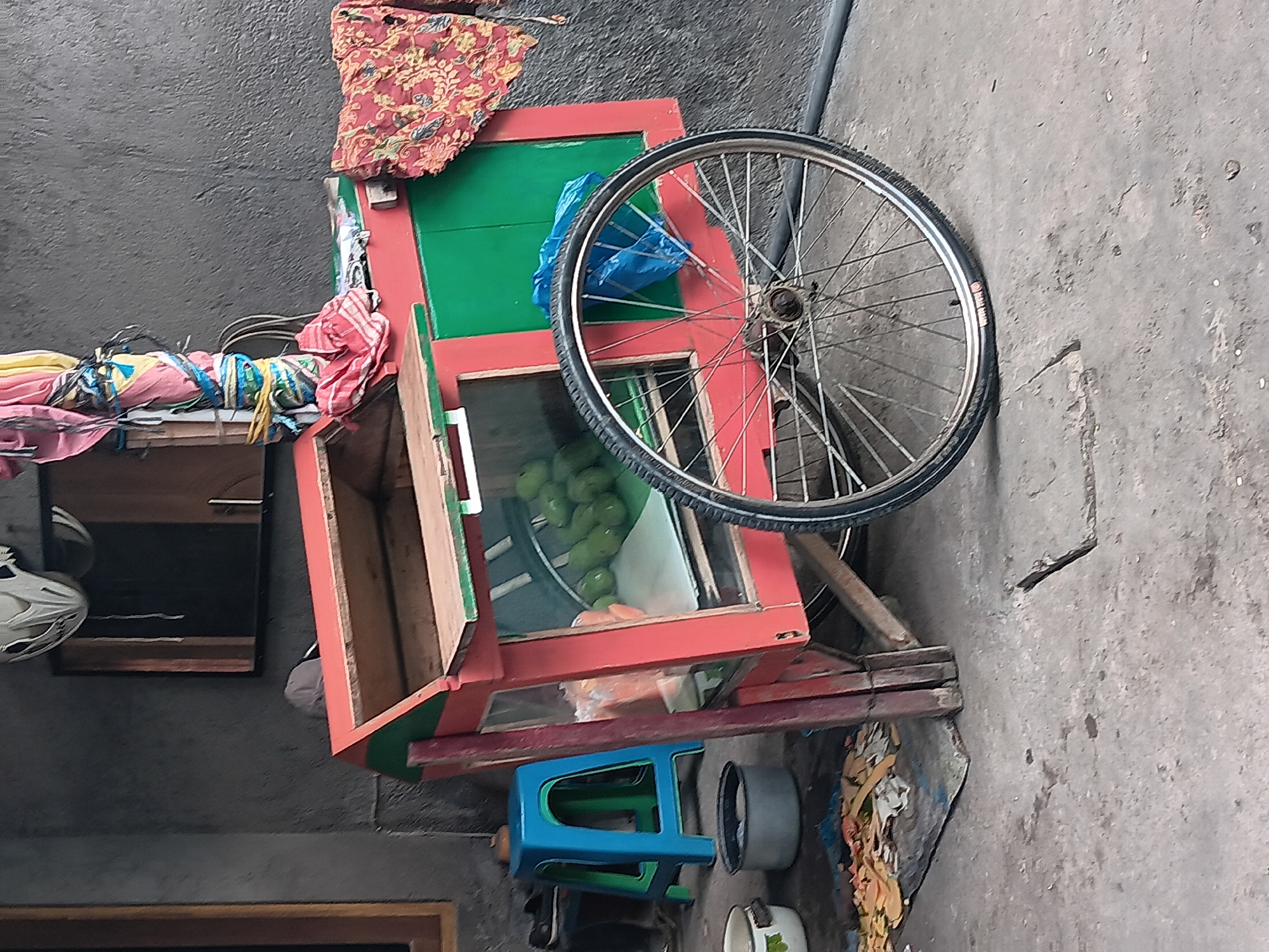 A wheeled cart with a red and green body, filled with green fruits, is parked on a concrete surface outside a building with a gray, textured wall in Batursari, Indonesia. The cart has a glass display case and a wooden counter. A bicycle wheel is attached to the front of the cart, and various items, including a blue plastic bag and a red patterned cloth, are draped on or near the cart. To the left of the cart, a blue plastic stool and two metal containers are visible. A white helmet hangs from the wall above the cart. The scene appears to be an outdoor stall or roadside vendor.