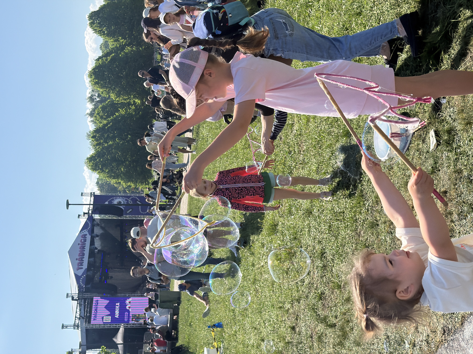 The image depicts an outdoor scene in Riga, Latvia, likely during a public event or festival. A stage with the word "TRADĪCIJA" is visible, as well as the name "ANNIKA" on a screen. A crowd of people is gathered in the background, and a young girl in a pink hat and dress is making large bubbles with a wand. Another child is also playing with bubbles. The setting appears to be a grassy area.
