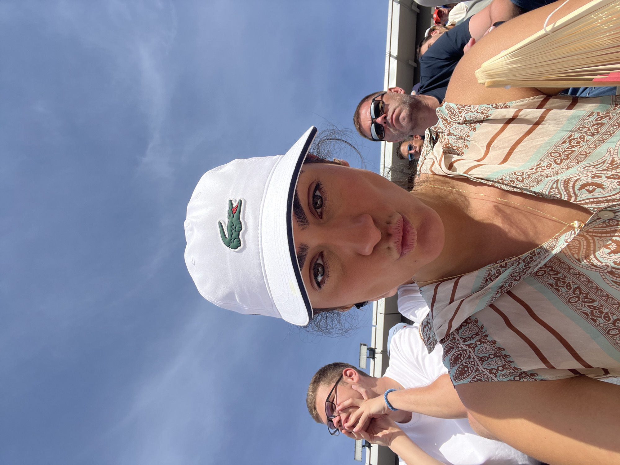A young woman is captured in a selfie, making a puckered lip expression while looking directly into the camera. She wears a white baseball cap with a prominent green Lacoste crocodile logo, and a patterned sleeveless top featuring stripes and paisley-like motifs in beige, brown, and light blue. A delicate gold chain with a small pendant is visible around her neck, and she holds a light-colored folding fan in her right hand.
To her left, a young man with short light hair and glasses wears a white t-shirt and a blue braided bracelet on his wrist; he clasps his hands near his chin, looking upwards. To her right, a man with stubble and dark sunglasses wears a dark t-shirt and looks forward with a serious expression. Other blurred spectators are partially visible behind them.
The scene is set outdoors under a clear blue sky with sparse wispy clouds, indicating bright sunshine. The individuals are seated in what appears to be stadium seating, with metal and white architectural structures visible in the background. Based on the Lacoste branding and the stadium environment, the location is identified as the Roland Garros tennis complex in Paris, France.