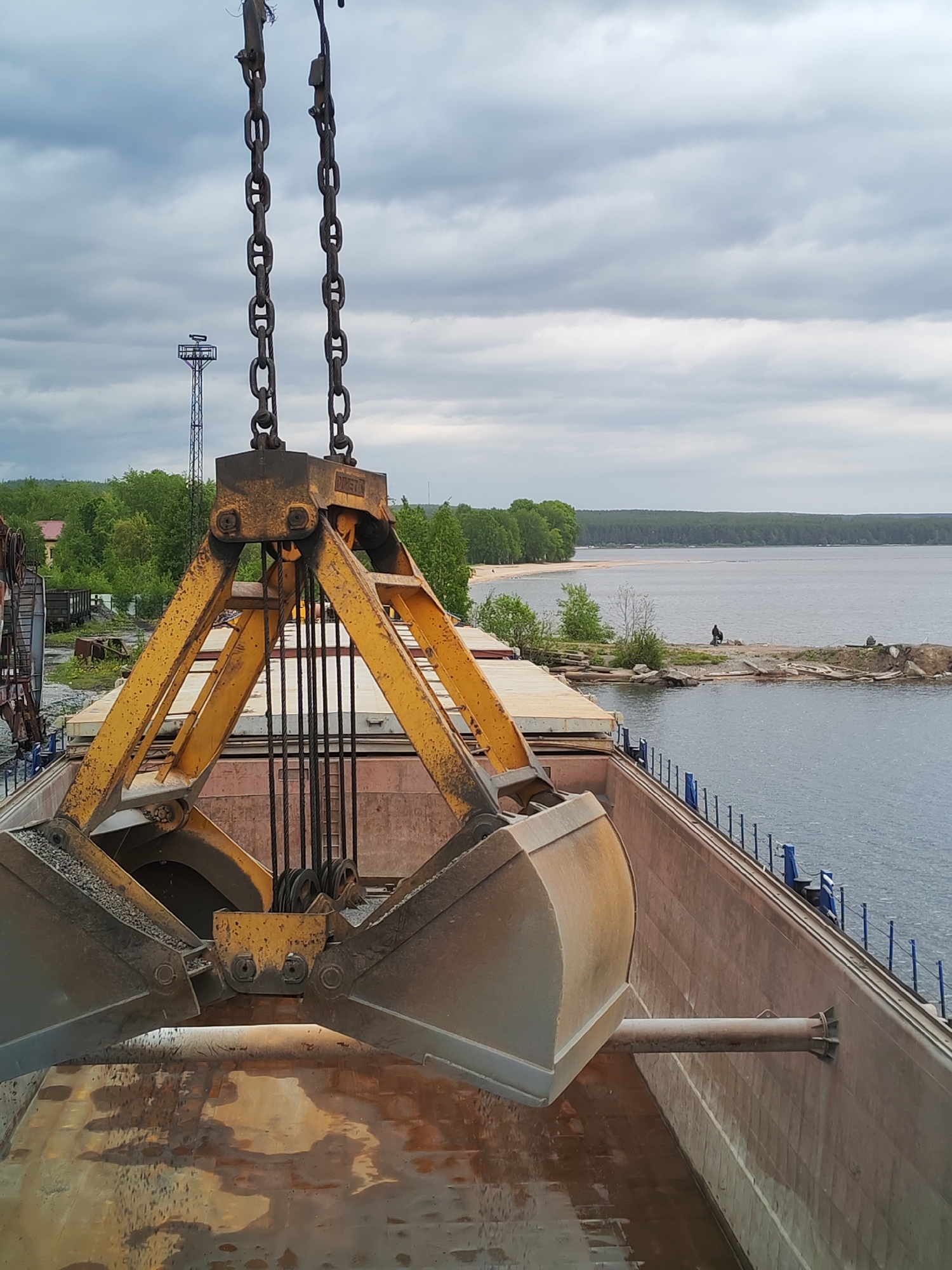 A large, yellow, industrial crane grab is prominently featured in the foreground, suspended above the open cargo hold of a ship or barge. The grab appears worn and rusty, partially open and containing what looks like gravel or other bulk material, indicating an ongoing loading or unloading operation. Heavy chains and cables connect the grab to an unseen crane mechanism above the frame.
The ship's rusty brown hull, equipped with blue railings and a horizontal support beam, extends into the right side of the image, floating on a large body of water. Across the water, a distant, tree-lined shoreline forms the horizon, featuring a sandy beach closer to the mid-ground. A small, rocky peninsula juts out into the water, where a solitary, dark-clad figure is seated.
In the left background, behind the crane structure, a tall, dark industrial tower or mast stands against the sky, with glimpses of green trees and potentially other industrial equipment or train cars further in the distance. The sky is overcast with heavy, gray clouds, suggesting a cloudy daytime setting. The scene depicts industrial activity at a port or dock, consistent with the location context of Medvezhyegorsk, Russia, likely on Lake Onega. Faint, partially obscured text is visible on the upper yellow structure of the crane grab, possibly reading "DIMMET" or similar.
