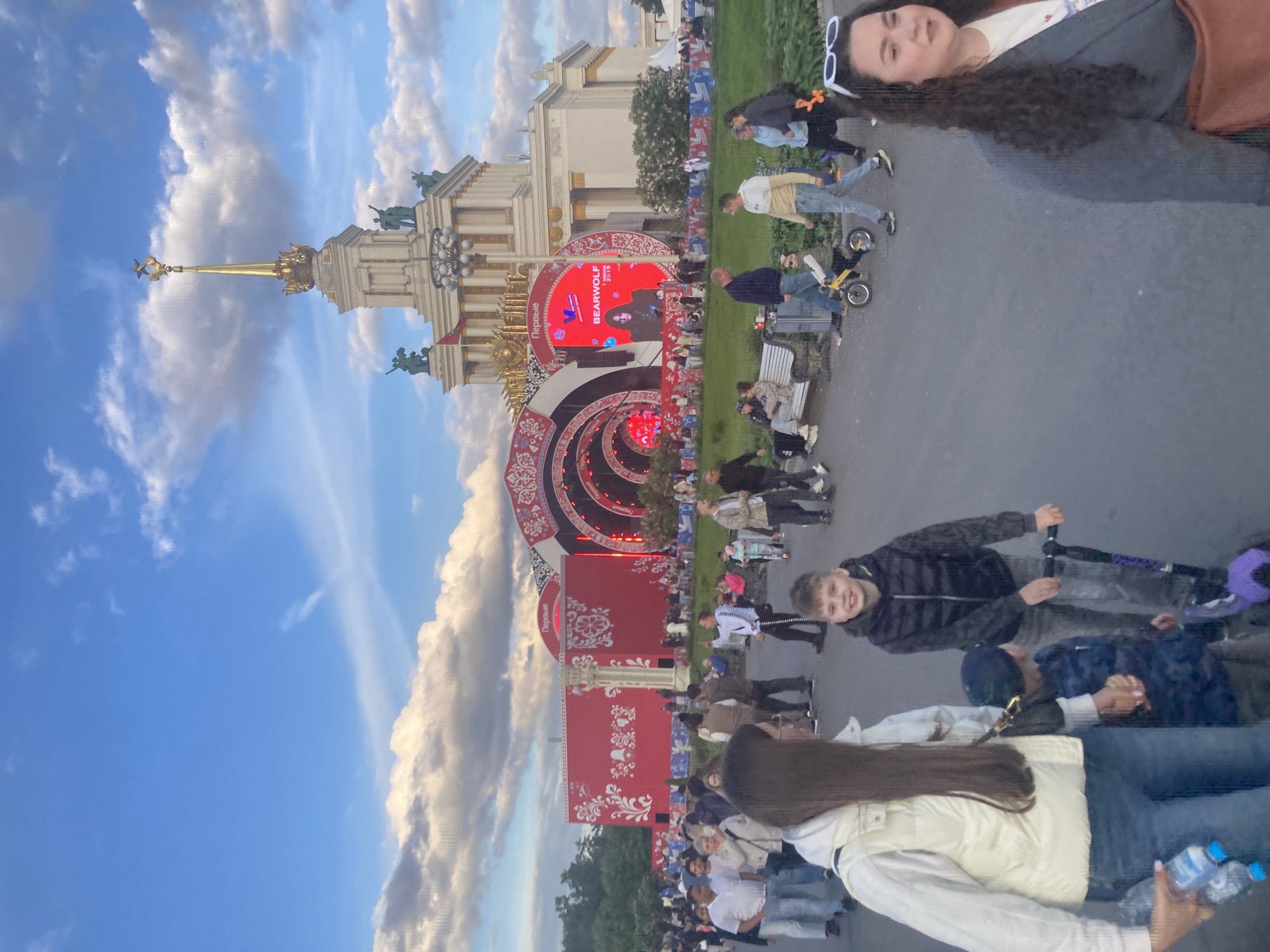 This image depicts a lively outdoor public event taking place at what appears to be VDNH (Vystavka Dostizheniy Narodnogo Khozyaystva) in Moscow, Russia, identifiable by the distinctive architecture. The scene unfolds in the late afternoon or early evening under a partly cloudy blue sky, indicating pleasant weather.
In the background, a grand, ornate building with a towering golden spire, crowned with a star and adorned with statues, dominates the skyline. In front of this architectural marvel, a large, elaborate stage in red and white, featuring traditional decorative patterns, is set up. A central circular screen on the stage displays the name "BEARWOLF" along with "2115" or "21:15," and the Russian word "Первые" (Pervye), meaning "The First" or "Pioneers." The screen also shows the image of a person, likely a performer or speaker.
A considerable crowd of people, comprising adults and children, is gathered in the open area before the stage. Many are standing and observing the stage, while others are seated on benches. The ground transitions between paved pathways and grassy sections.
In the foreground, several individuals are visible. On the right side of the frame, a woman with long, dark, curly hair and white sunglasses on her head looks towards the camera. Closer to the center, a young boy on a scooter smiles at the camera, dressed in a dark jacket. Towards the bottom edge, another woman with long dark hair, wearing a white jacket, is seen holding two plastic water bottles. The overall atmosphere appears relaxed and festive, suggesting a concert or public festival is underway.