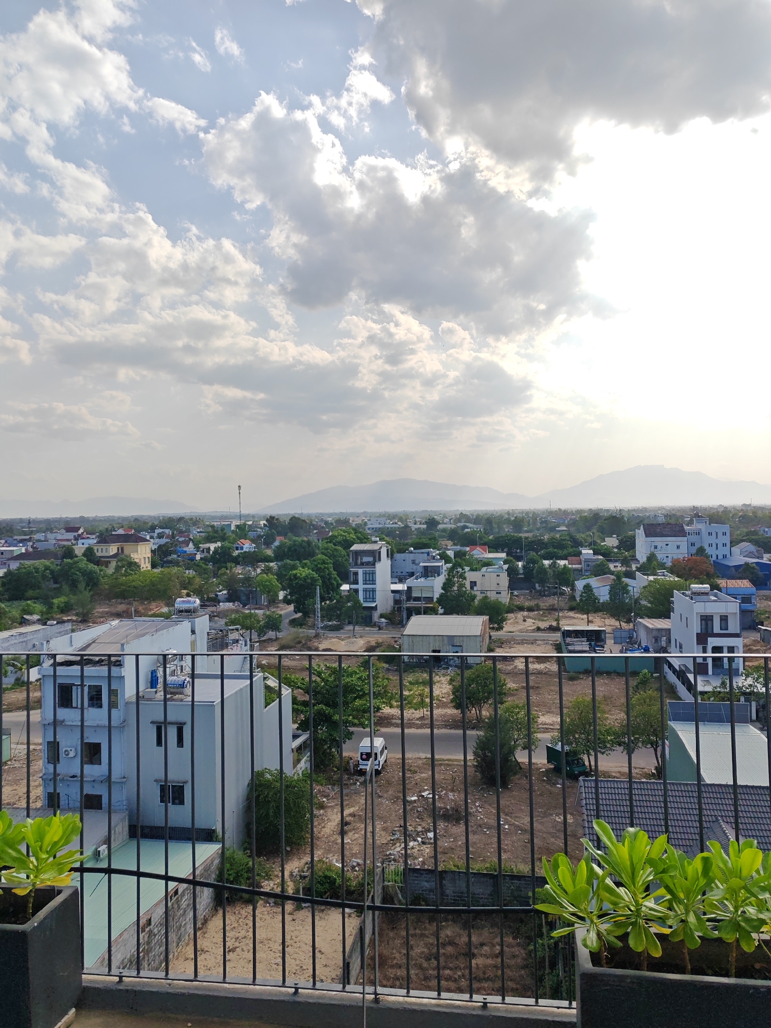 The image captures an elevated view of a city with a hazy sky, likely in Đà Nẵng, Vietnam. In the foreground, a black metal railing borders a balcony, with two potted plants containing bright green foliage visible on the left and right edges. Beyond the railing, a street and several buildings are visible. A light blue, three-story building with numerous windows and air conditioning units stands out on the left. Across the street, a white van is parked. Further into the cityscape, a mix of residential and commercial buildings, some with flat roofs and others with tiled roofs, are scattered amidst lush green trees. In the distance, rolling hills or mountains form a hazy horizon under a bright, cloudy sky. The overall impression is of a developing urban area with a mix of modern architecture and natural greenery.