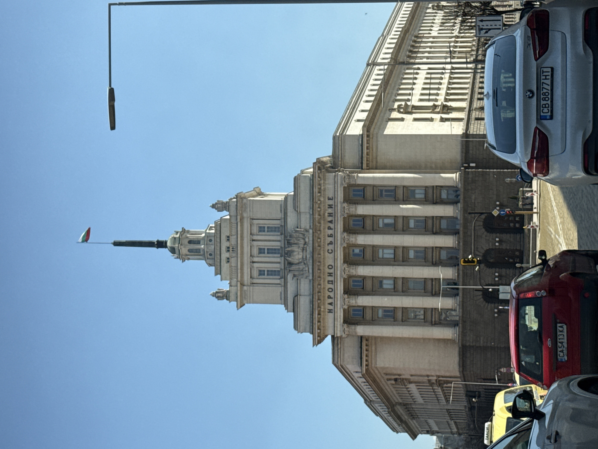 The image captures a view of the National Assembly of Bulgaria (Народно събрание) in Sofia, Bulgaria. The building is a light-colored, classical-style structure with a prominent spire topped by a Bulgarian flag. The inscription "НАРОДНО СЪБРАНИЕ" is visible on the building's facade. The sky is clear and blue. In the foreground, there is a street scene with several cars parked or moving. One car is a white BMW X5 with license plate "CB 8877 HT," and another car is red with license plate "CA 4113 KA." There is a street lamp on the left side of the image. The photo appears to have been taken from street level, looking upwards towards the building.