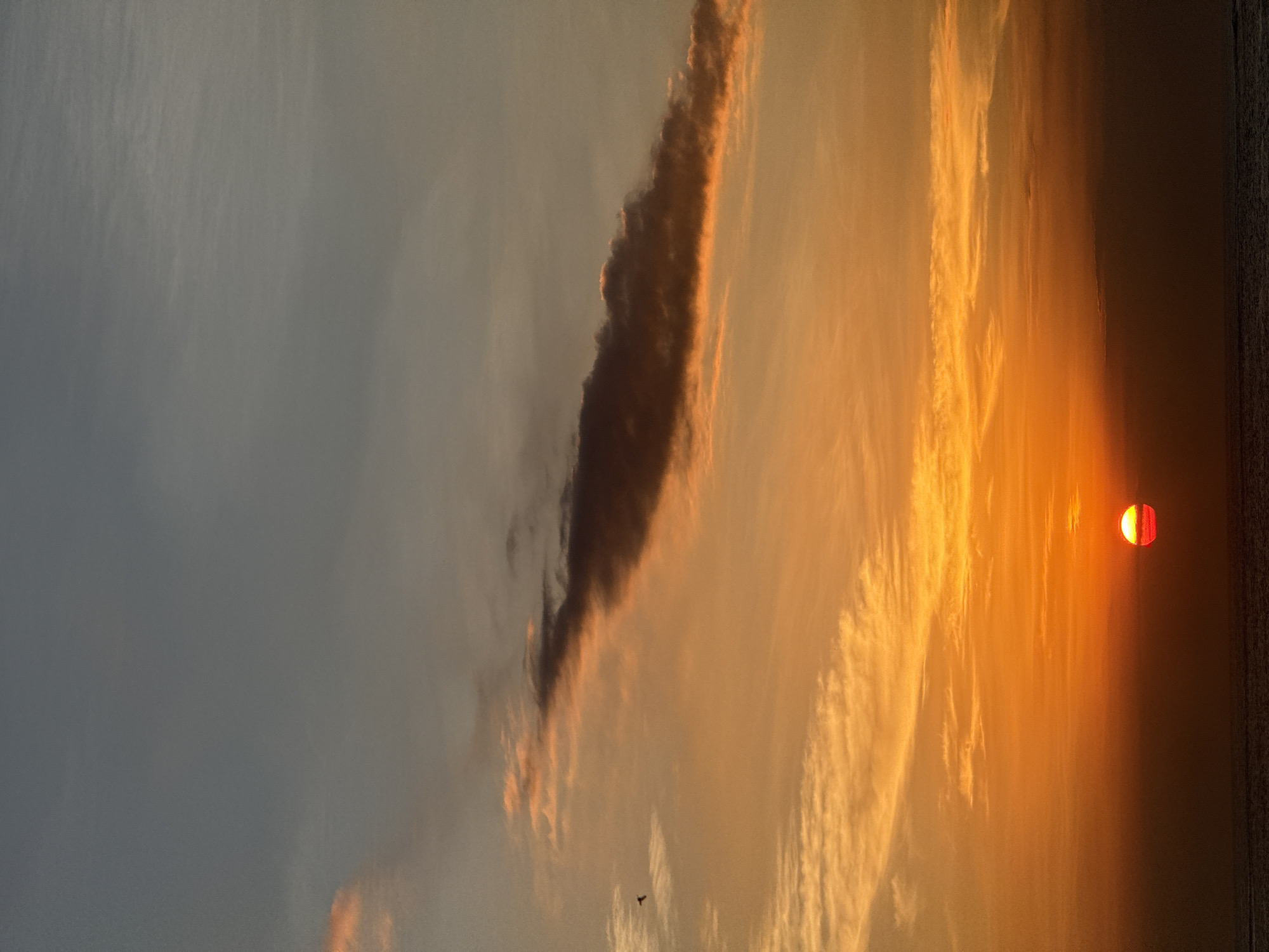 The image depicts a vibrant sunset over the ocean in North Kuta, Indonesia. The sky is filled with dramatic clouds, ranging from dark, ominous formations to wispy streaks illuminated by the setting sun. The sun itself is a bright, reddish-orange orb partially obscured by the horizon, casting a warm glow across the scene. The ocean is visible as a dark band at the bottom of the frame, with its surface texture suggesting gentle waves. The overall atmosphere is one of awe and tranquility, as the natural spectacle unfolds. The time of day is clearly sunset, with the intense colors of the sky indicating the sun's descent. There are no people or man-made objects visible, emphasizing the natural beauty of the landscape. The image captures a moment of serene beauty, evoking a sense of peace and wonder associated with sunsets. There is no visible text in the image.