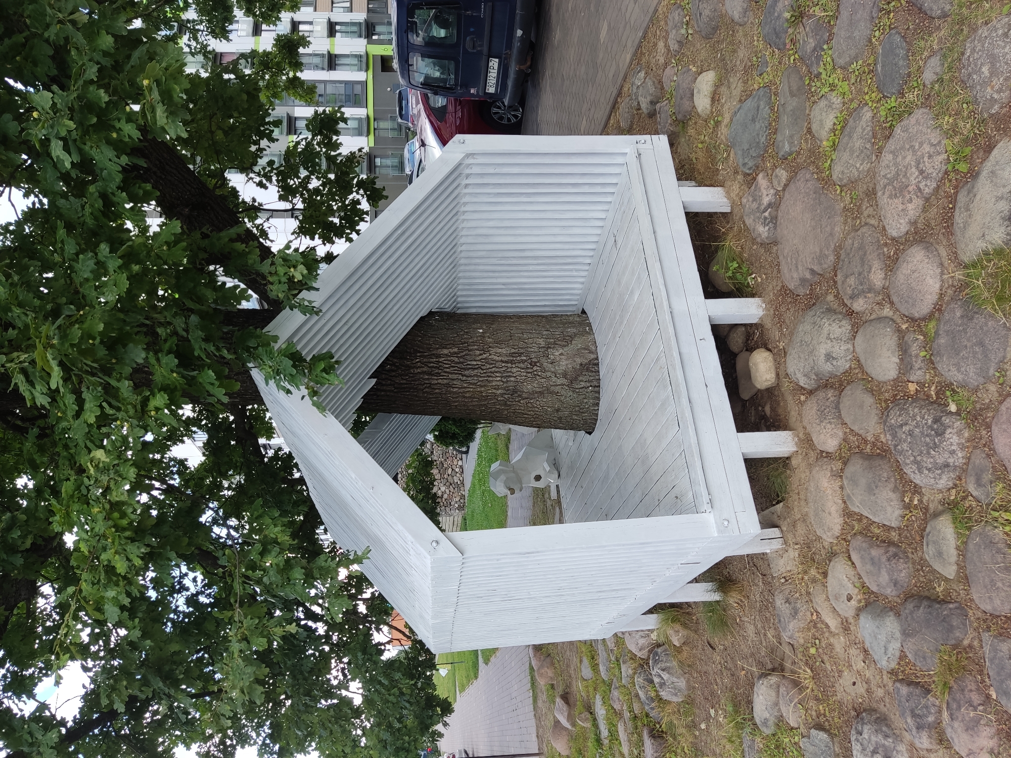 The image captures a unique outdoor installation in the city of Бараўлянскі сельскі Савет, Belarus. At the center, a large tree trunk emerges from the floor of a white, open-sided structure with a corrugated metal roof. The structure has a wooden floor and walls composed of vertical planks, painted white. Inside the structure, to the left of the tree, sits a stylized, geometric white sculpture of a dog. The installation is situated on a ground surface covered with a mix of cobblestones, smaller rocks, and patches of grass. In the background, a modern apartment building with a green and white facade is visible, along with parked vehicles, including a dark blue van with a license plate reading "8212 TP-7". A paved walkway runs along the left side of the image. The scene appears to be outdoors in an urban or suburban setting.