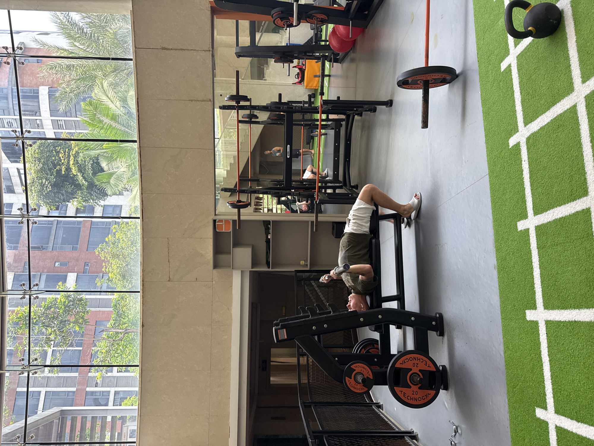 The image depicts an indoor fitness center in the Haizhu District, China. In the foreground, a male subject is positioned supine on a black weight bench, actively performing a barbell press with a metal barbell. He wears a dark olive green t-shirt, white shorts, and white slip-on sandals. Adjacent to the bench, black and orange Technogym weight plates, some marked "20 KG," are visible on the equipment rack and the floor. The flooring consists of a gray polished concrete section, transitioning to a green artificial turf area with white grid lines in the lower portion of the frame.
A large black squat rack with orange safety bars is centrally located in the midground, holding multiple barbells. Behind this rack, a substantial mirror wall reflects the interior. Within the reflection, a second male subject stands upright, holding a bottle, attired in a dark sleeveless top and light shorts. The lower body of a third individual is also partially visible in the reflection near the rack. To the right, two red exercise balls are stacked, and a black barbell with a single orange and black weight plate lies on the green turf. A black kettlebell with a yellow stripe is positioned on the turf in the lower right corner.
The upper section of the gym is characterized by a light beige tiled wall that seamlessly integrates with expansive, floor-to-ceiling glass windows. These windows provide an unobstructed view of an exterior urban landscape, featuring multi-story buildings with red brick facades and numerous windows, interspersed with dense green foliage, including trees and prominent palm trees. The sky overhead appears bright. A dark metal mesh fence borders the left side of the gym interior.