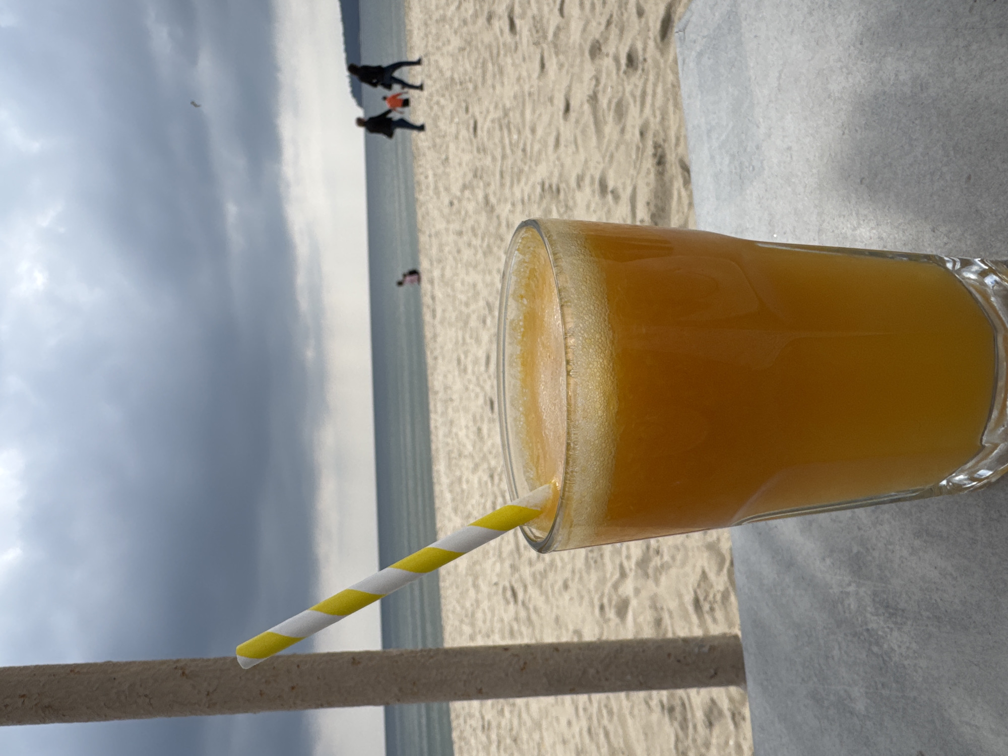 A glass of orange juice with a yellow-and-white striped straw rests on an outdoor table. The background shows a sandy beach with a few people walking and the sea under a cloudy sky. The image may be taken on the beach in Varna, Bulgaria.