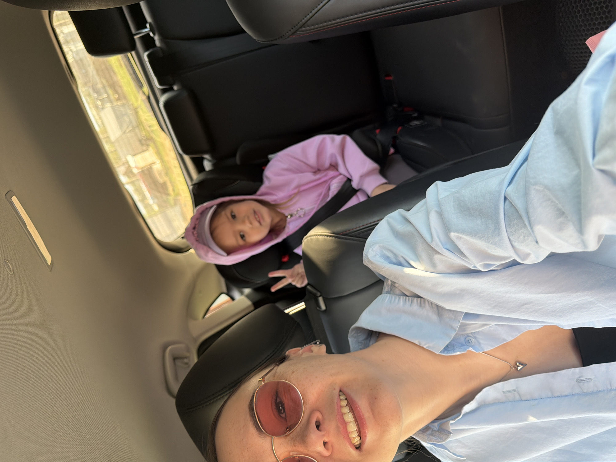 From inside a car, a young girl wearing a pink hooded shirt and fastened in a car seat gestures with her fingers. A woman in a light blue shirt and rose-colored sunglasses is taking the photograph. The location appears to be inside a vehicle, but there are no specific details in the image to ascertain whether the location is in Chaikovsky, Russia.
