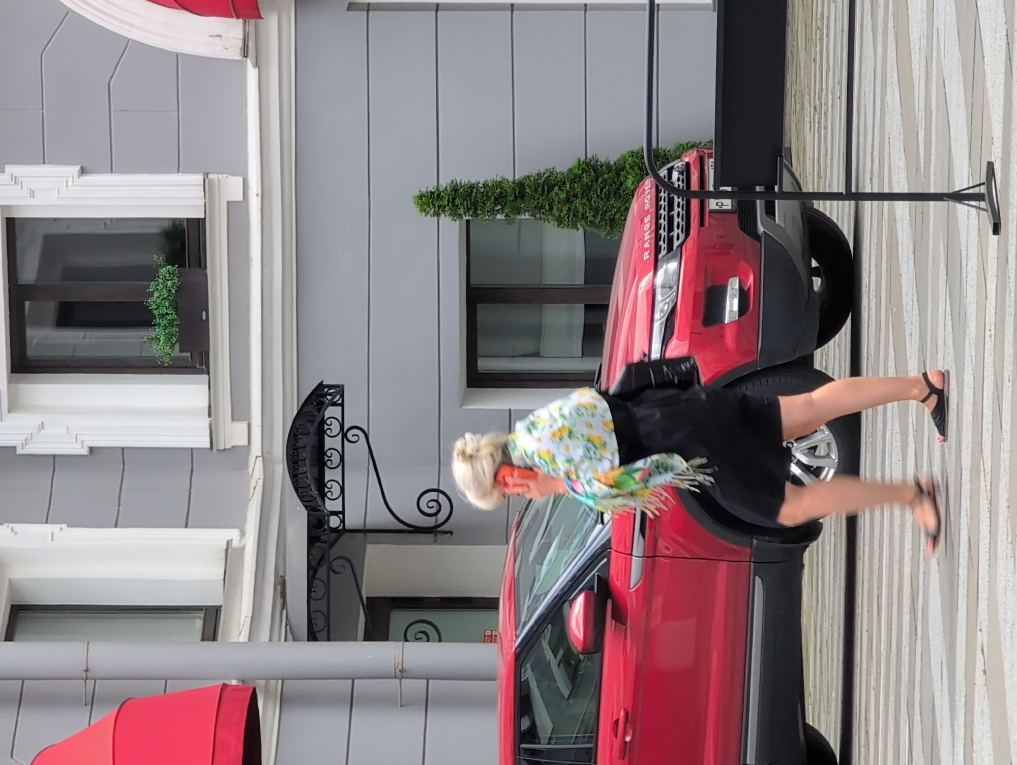 A blonde woman in a black dress and a colorful scarf is walking across a crosswalk in front of a red Range Rover on a cloudy day. She is holding a phone to her ear. The building behind her is grey with white trim. A tall green bush is visible on the right side of the image. The scene appears to be in an urban setting, possibly in Saint Petersburg, Russia, given the architectural style and presence of a Range Rover.