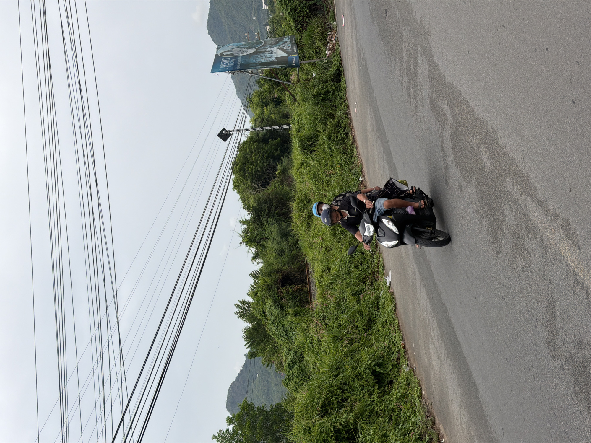 An overcast day in Nha Trang, Vietnam, shows two individuals riding a black scooter down a paved road. The person in front, wearing a black shirt and a blue helmet, steers the scooter. Behind them, a second person, dressed in a black patterned garment, sits as a passenger. Lush green vegetation lines the side of the road, with a billboard for "I-RESORT" visible on the right. In the distance, a mountainous landscape under a gray sky suggests the outskirts of the city. Power lines crisscross overhead.
