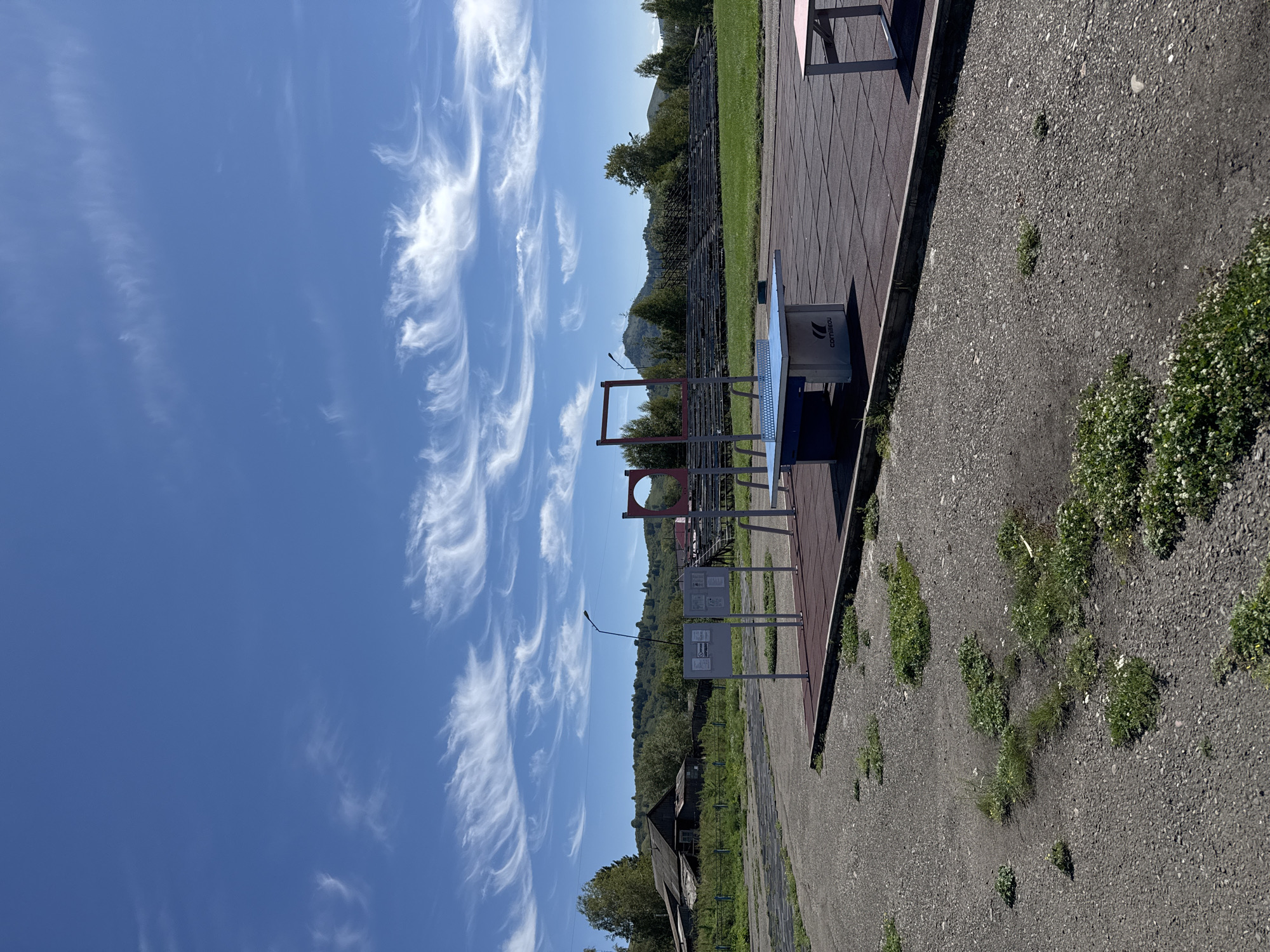 This outdoor image shows a public recreational area with a ping pong table and exercise equipment under a bright blue sky with wispy cirrus clouds. In the foreground, a ping pong table is set on a red rubberized surface, with another table visible to its right. Behind the ping pong table, there are red frames of what appear to be exercise apparatus. Further in the background, there are bleachers for spectators, a wooded hill, and an old wooden building with a metal fence. The area appears to be located in Soloneshnoye, Russia, given the context of the image. The ground is a mix of gravel and some sparse vegetation.