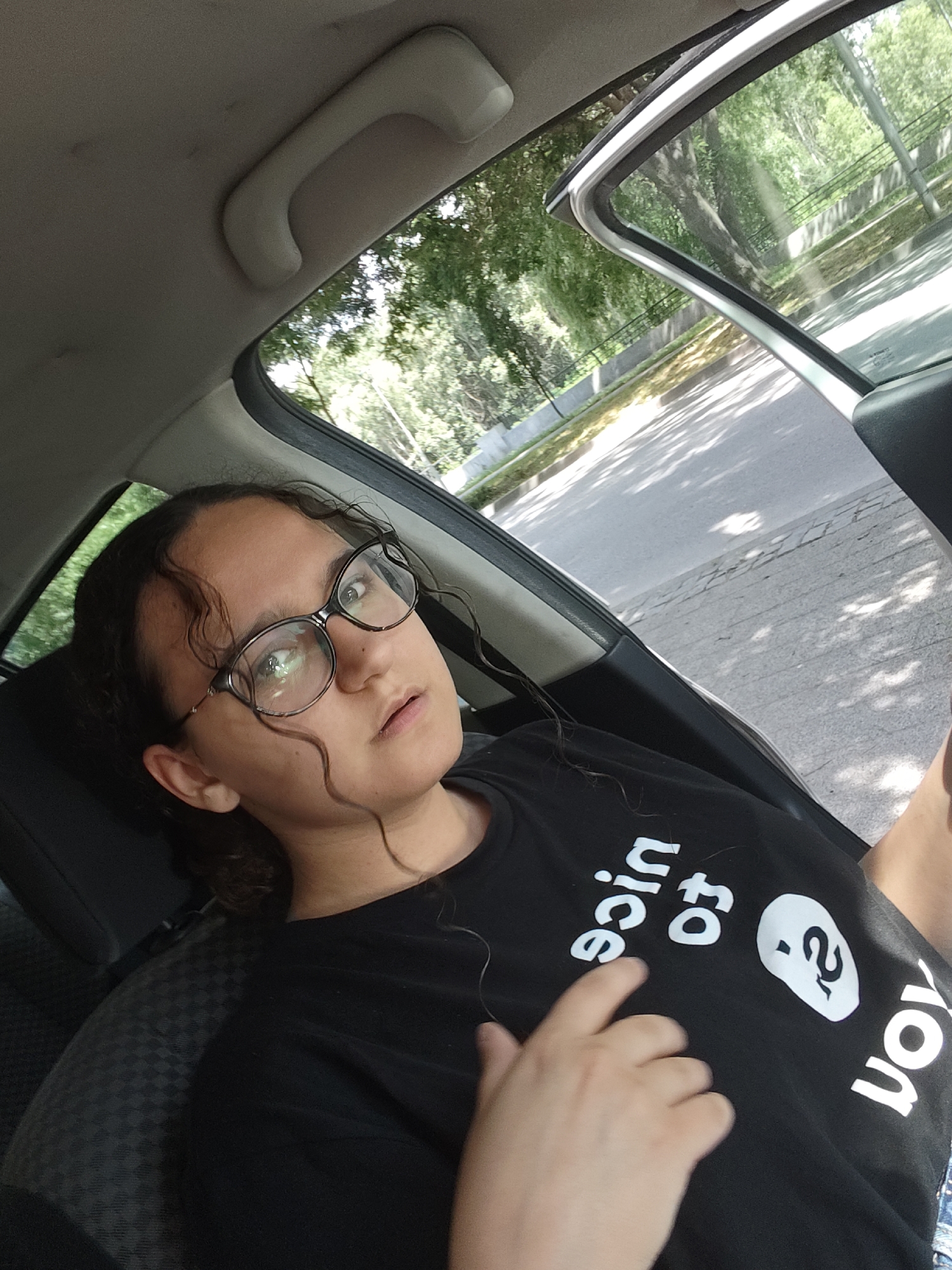 A young woman with dark curly hair and glasses is sitting in the passenger seat of a car. She is wearing a black t-shirt with white text that reads "nice to..." and a stylized drawing of a face. The car door is open, and the woman is looking slightly off-camera with her mouth slightly open. Outside the car, trees and a road are visible. The image appears to be taken during the day, with sunlight casting shadows.