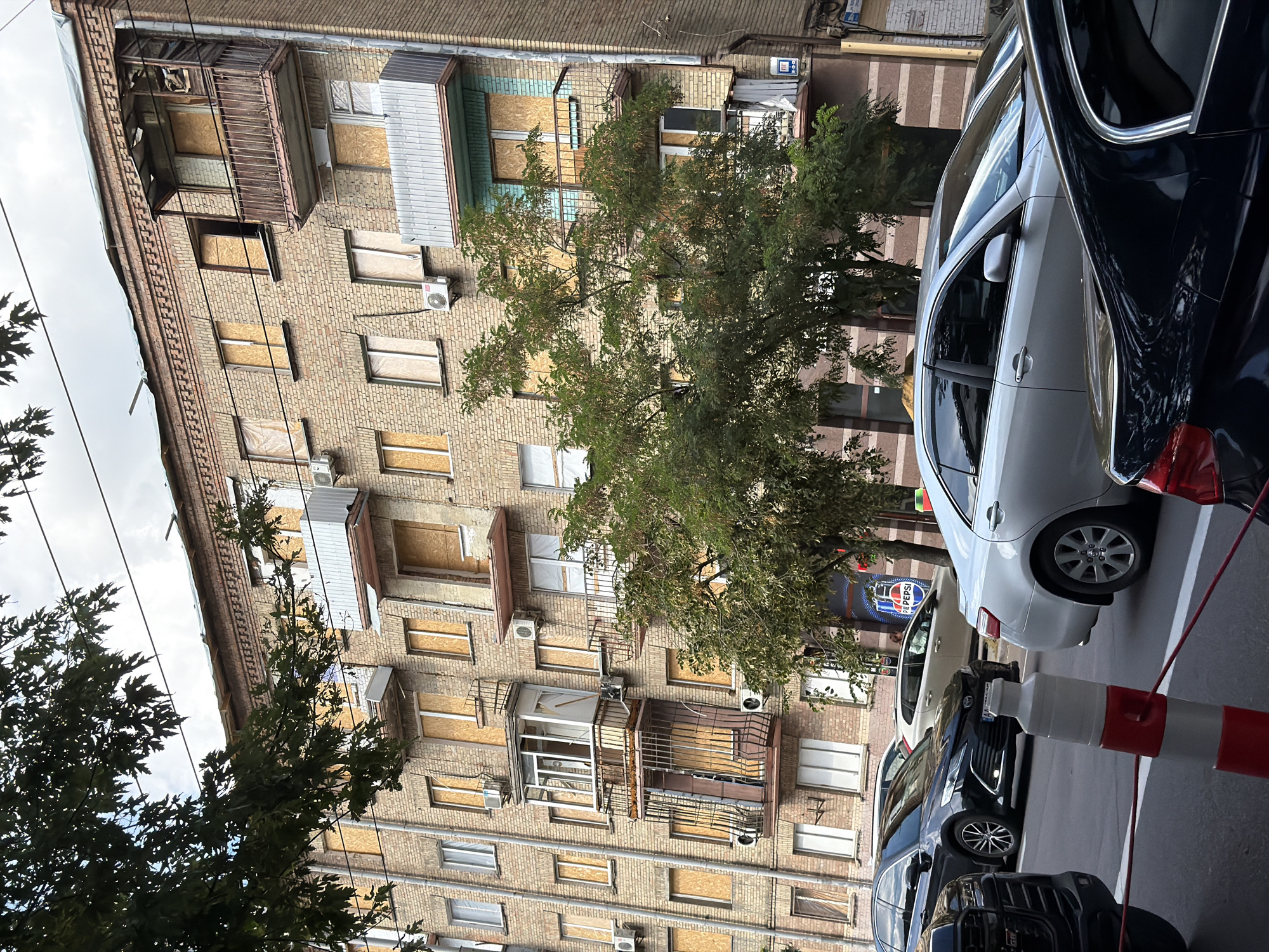 This is a street-level, medium shot of a multi-story brick apartment building in Kyiv, Ukraine, with several windows and balconies boarded up. The building appears damaged, with some sections of the roof also covered with tarps. Trees with green foliage are in the foreground, partially obscuring the lower levels of the building. Several cars are parked on the street in front of the building. One car is silver, another is black, and a third car is visible in the background. A traffic cone with red and white stripes is in the foreground, with red cords extending from it. The sky is overcast.