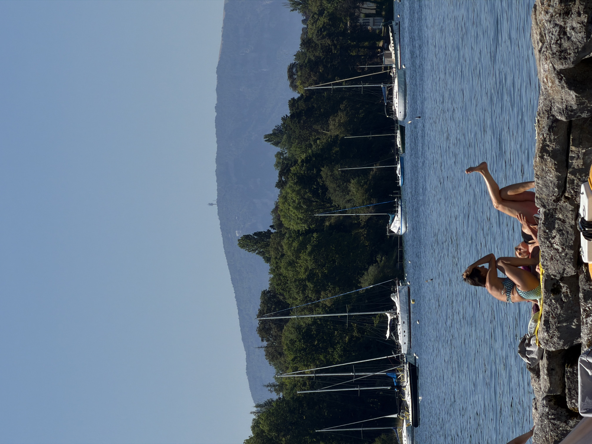This image captures a sunny day at the lake, likely in or near Hermance, Switzerland, with the Alps in the background. In the foreground, two individuals are relaxing on a rocky shoreline. One person is sitting upright, wearing a patterned bikini, with their hand resting on their head. The other person is lying down, with their legs extended and slightly bent, and is wearing a dark bikini top and a tan bottom. A yellow towel and a gray bag are placed near them on the rocks.
The middle ground is dominated by a body of water, likely Lake Geneva, dotted with numerous sailboats. The sailboats are of various sizes and designs, with their masts tall against the blue sky. Some are moored, while others appear to be anchored. The water is a calm blue, with subtle ripples.
The background features a dense line of lush green trees bordering the lake, suggesting a park or forested area. Beyond the trees, a large, hazy mountain range, possibly the Alps, forms the horizon. A prominent telecommunications tower is visible on one of the mountain peaks. The sky is a clear, bright blue, indicating fair weather. The overall atmosphere is one of leisure and tranquility, typical of a summer day by the water.