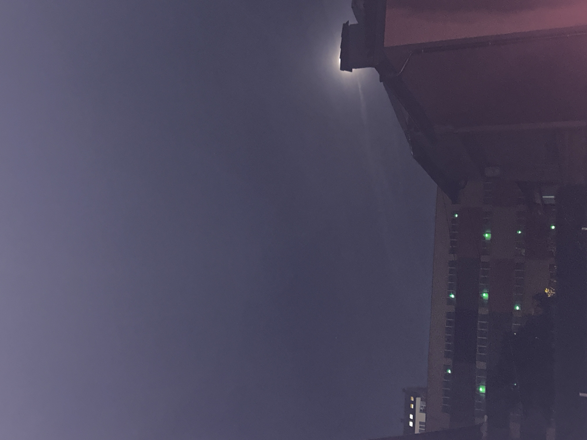 Captured at night, the image reveals a section of Sofia, Bulgaria. The sky, a muted purple, dominates the frame. A single light source, likely a street lamp, shines brightly near the upper edge of the image, partially obscured by an overhanging structure. A building rises in the right portion of the image, its height suggested by the repeating pattern of illuminated windows, some glowing with a green hue. The composition is vertical, focusing on the upward view, and the lighting implies it is after dusk.
