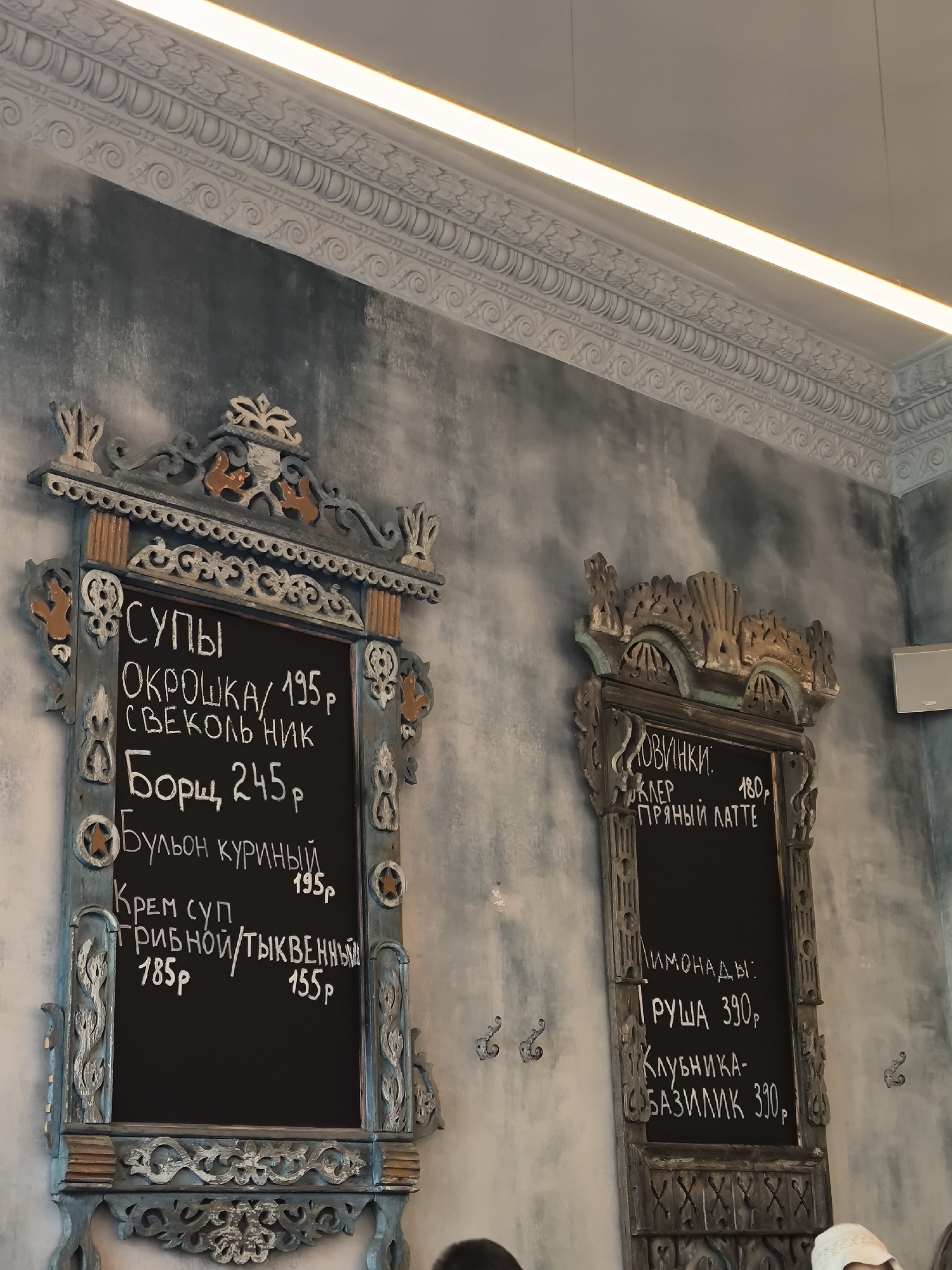 The image captures a scene indoors, likely in a restaurant or café in Unknown, Unknown. The primary focus is on two ornately framed menus hanging on a textured, grey-blue wall. The frame on the left, painted in blue and orange, displays a menu of soups written in Russian. The menu lists items such as "ОКРОШКА" (Okroshka), "СВЕКОЛЬНИК" (Beetroot Soup), "Борщ" (Borscht), "Бульон куриный" (Chicken Broth), and cream soup varieties. Each item is accompanied by a price in Russian rubles, "р". The second menu, on the right, also features a decorative frame and displays a list of beverages, including "Пряный латте" (Spicy Latte) and lemonades made with pear ("ГРУША") and strawberry-basil ("Клубника-БАЗИЛИК").
The top edge of the wall is embellished with ornate, white plaster moldings that run along the ceiling, beneath a strip lighting fixture. Partial views of people can be seen at the bottom of the frame, and there's a wall-mounted speaker visible on the right side of the frame. The overall lighting appears to be a combination of artificial light from the ceiling fixture and ambient light, casting shadows on the wall.