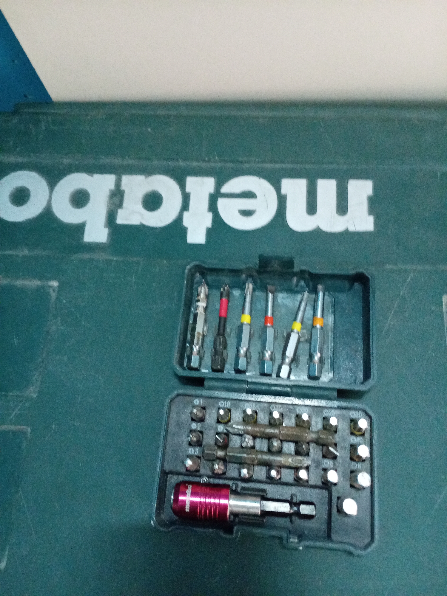 The image shows a Metabo brand toolbox with a set of screwdriver bits inside. The toolbox is green with the word "metabo" written in white capital letters. The screwdriver bit set is in a black plastic case, which is open to reveal an assortment of various sized and shaped bits, including torx, flathead, and Phillips. One bit has a red anodized aluminum holder. The background is a plain, light-colored surface, possibly a wall or table.