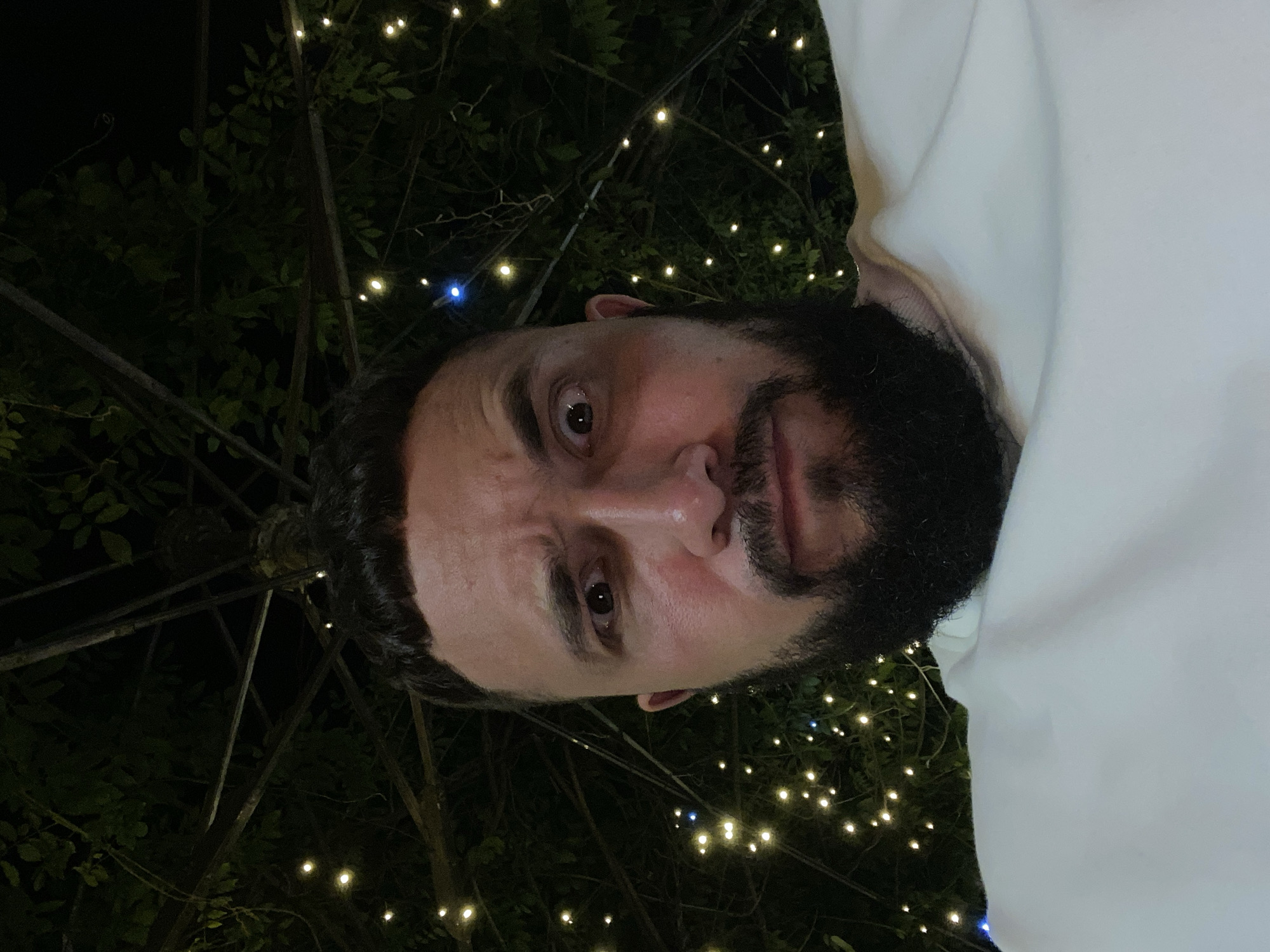 A close-up, low-angle selfie of a man with dark hair and a full beard is taken outdoors at night. He is wearing a white sweatshirt. The background is dark with blurred, twinkling fairy lights and foliage, suggesting a tree canopy or trellis. The man's expression appears surprised or shocked, with wide-open eyes and furrowed brows. The image does not provide any information about the city of Makhachkala, Russia, nor any specific location within it.