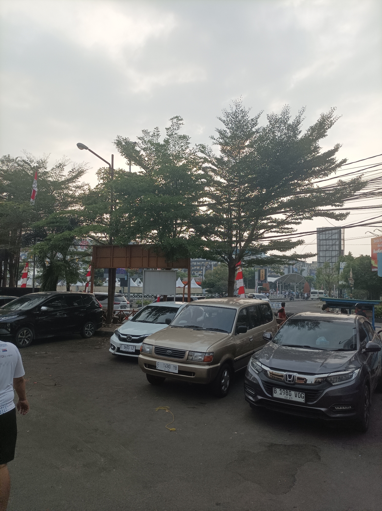 The image depicts an outdoor scene with several cars parked on a paved surface. In the foreground, a dark gray SUV is prominently displayed, facing the viewer. To its left, a beige SUV is parked, also facing forward. Behind these vehicles, a white sedan is visible. Further back, a black SUV is parked on the left side of the frame.
The environment features several trees with dense foliage, some of which are illuminated by a soft, overcast sky. In the background, there are buildings and utility poles with numerous wires. Red and white flags are displayed on poles along the left side of the image. The lighting suggests it is daytime, possibly late afternoon or early morning, due to the diffused light. The scene appears to be a parking lot or an area adjacent to a road, likely in an urban setting.
Specific details from the image and provided crops suggest the location is in Cirebon, Indonesia. The license plate on the dark gray SUV reads "B 1980 VOG". The license plate on the white sedan reads "E 1633 LT". The license plate on the beige SUV reads "E 1480 YB". The presence of "B" on one license plate suggests Jakarta, but "E" plates are typically from West Java, including Cirebon. The general urban landscape with the architectural style of buildings and the types of vehicles are consistent with an Indonesian city.