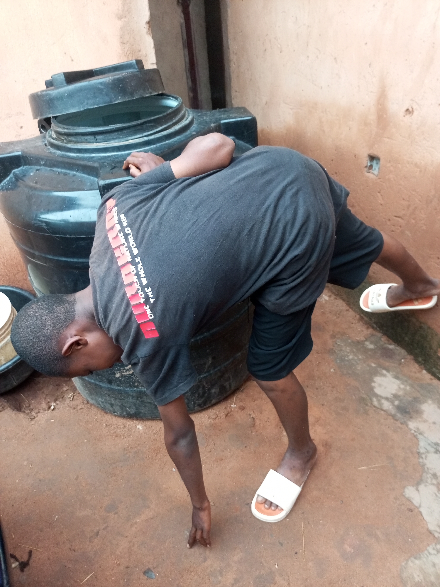 An eye-level shot shows a person bending over in front of a large black water tank. The person is wearing a dark grey t-shirt with red lettering that reads "ONE TOUCH OF NATURE THE WHOLE WORLD HINS". They are also wearing dark shorts and white slide sandals on their feet. The person's head is down, and they appear to be reaching into a black basin next to the water tank. The ground is a reddish-brown dirt, and the wall behind the water tank is a pale orange color. The image does not provide enough information to determine the location in the city of Tempure, Nigeria.
