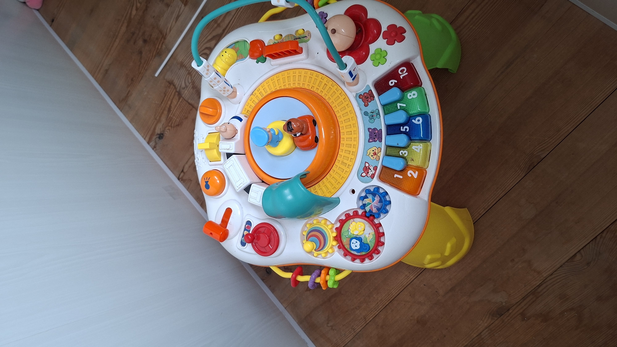 This is a top-down, medium shot of a colorful, plastic baby activity toy resting on a wooden floor. The toy is white with various interactive elements in bright colors, including orange, green, blue, yellow, and red. A teal archway is visible at the top, with a yellow and orange keyboard section to the right. The keyboard has blue keys with numbers and colorful animal graphics on the white surface.
In the center of the toy is a circular spinning platform with a light blue background and a cartoonish dog figure with a blue hat. There are other interactive features such as orange buttons, red knobs, and spinning gears with animal designs. A yellow plastic piece juts out from the bottom right of the toy.
The setting appears to be indoors, with a light-colored wall on the left and a brown wooden floor with visible planks and grain patterns. The lighting suggests it is daytime, with no strong shadows. No people are visible in the image. The overall mood is playful and child-oriented due to the toy's design.
There is some text on the toy, visible on the keyboard area and on some of the interactive buttons, but it is too small to be legible. The location context provided is Chaikovsky, Russia, but there are no specific visual cues within the image to confirm this.