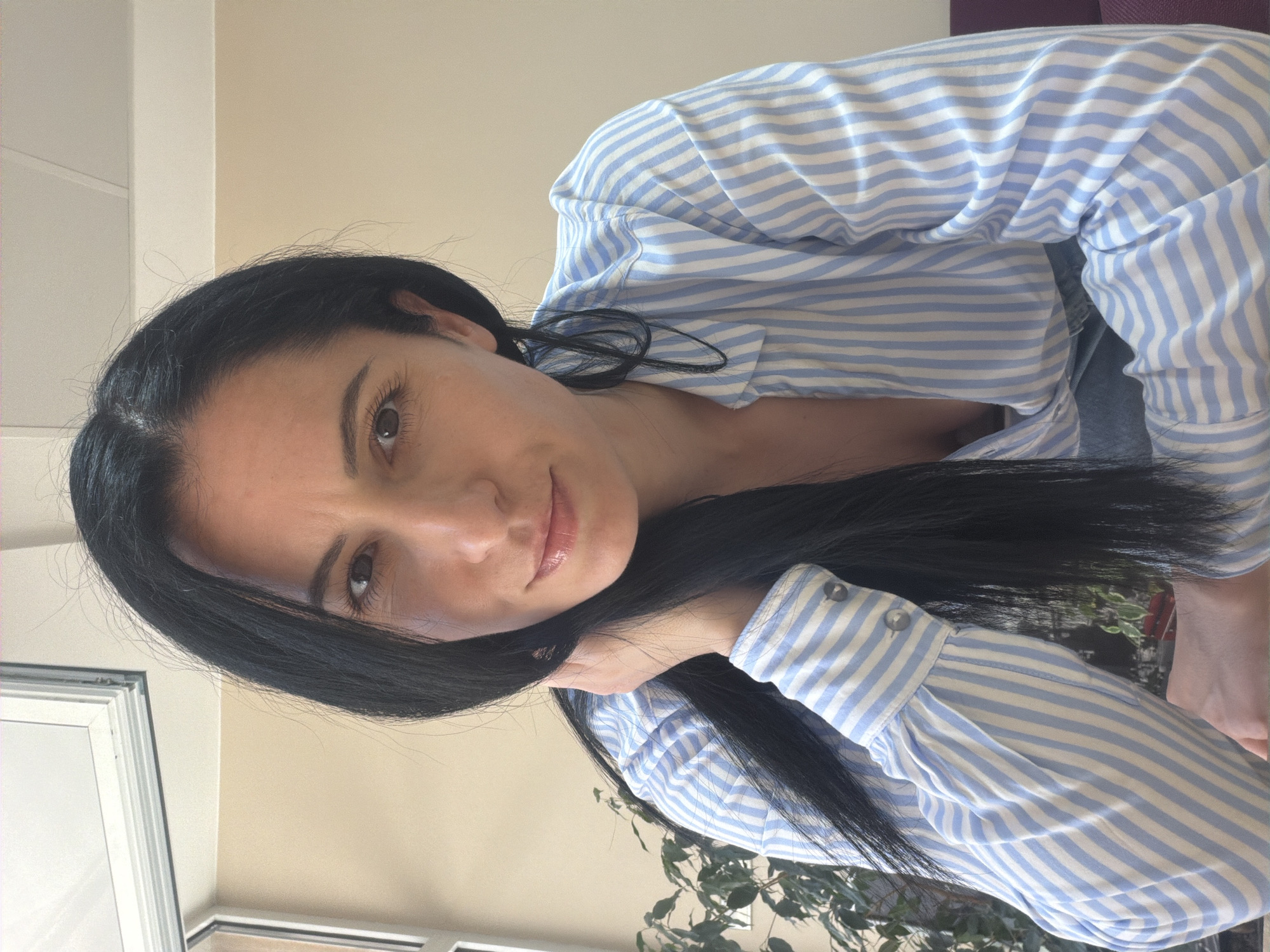 A woman with long, dark hair and light skin is looking at the camera. She is wearing a blue and white striped shirt with buttoned sleeves. Her left hand is resting near her neck. Behind her, a plant with green leaves is visible. The background consists of neutral-colored walls and a window. There is nothing in the image to suggest that this image was taken in Pleven, Bulgaria.