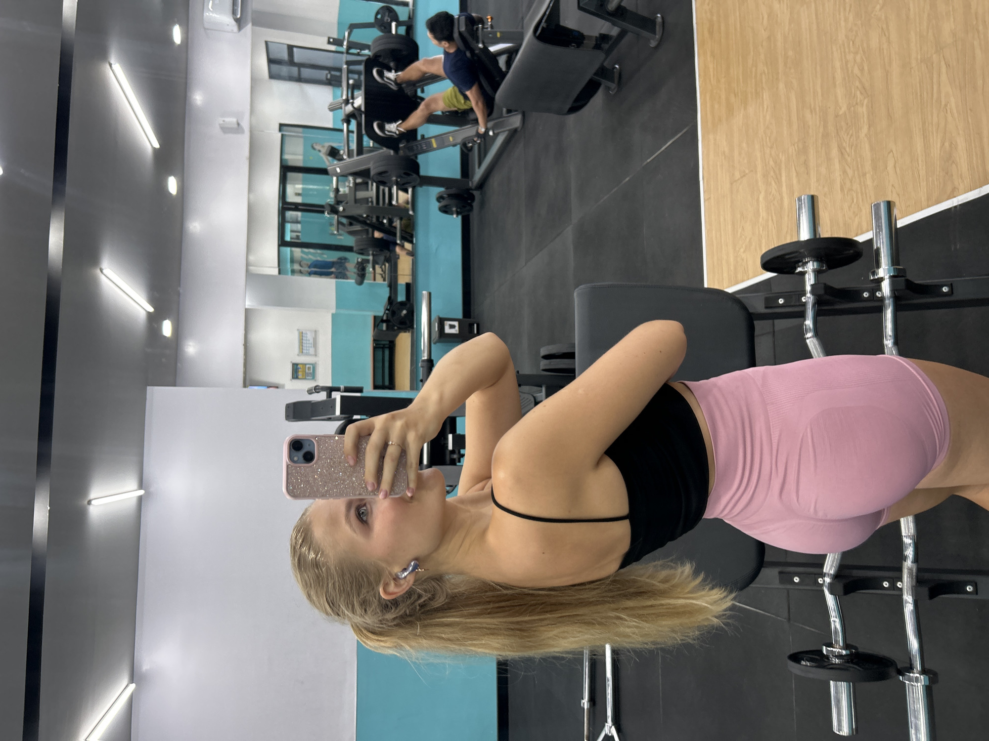 A young woman stands in a fitness center in Kuta Selatan, Indonesia. She is taking a selfie with a pink cell phone; she wears a black tank top and pink athletic shorts. Her blonde hair is pulled back, and she wears blue earrings. To her right, a weight rack holding plates stands. In the background, exercise equipment is visible and another person is using equipment. The room has mirrors, fluorescent lights, and blue and white accent walls. The floor is dark gray, with a wood-paneled wall in the upper right.