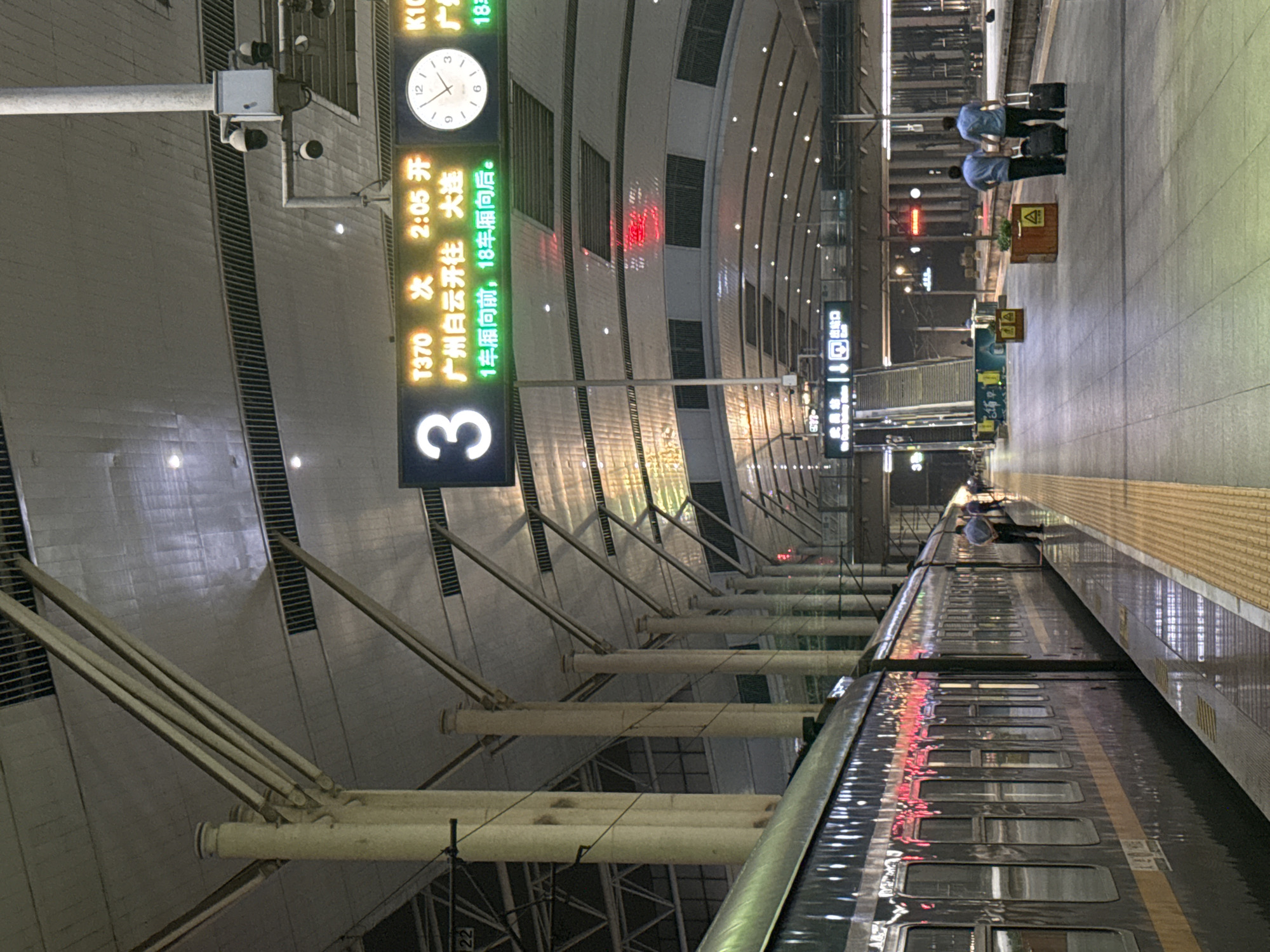 The image depicts a train station platform in Wuchang District, China. A dark-colored train is parked alongside the platform, its windows reflecting red and white lights. The platform itself is paved with gray tiles and features a yellow safety strip along the edge. Overhead, a large, illuminated sign displays train departure information, including train numbers, destinations, and times. The sign indicates train T370 departing at 2:05 for Dalian, and train K102 at 18:xx, also for Dalian. A circular clock above the departure information shows the time as approximately 12:09.
Several individuals are visible on the platform. Two men in light blue shirts and dark pants are walking away from the camera, each pulling a black suitcase. Another person, wearing a dark shirt and pants, stands near the train, looking in the direction of the camera. In the background, a set of escalators leads up to a higher level of the station. The station ceiling is a large, arched structure supported by white pillars, with lighting fixtures visible. The overall scene suggests an evening or nighttime setting within a modern, well-lit train station.