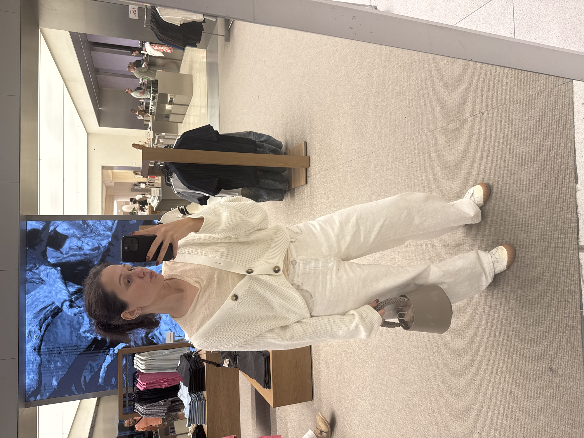 A person is standing in what appears to be a clothing store, taking a picture of themselves in a mirror. The person is wearing a white cardigan, a light-colored shirt, white pants, and sneakers. They are holding a small, round handbag. The store has a modern interior with clothes racks and displays. In the background, other shoppers can be seen browsing. The scene suggests a retail environment, possibly in Dubai, UAE.