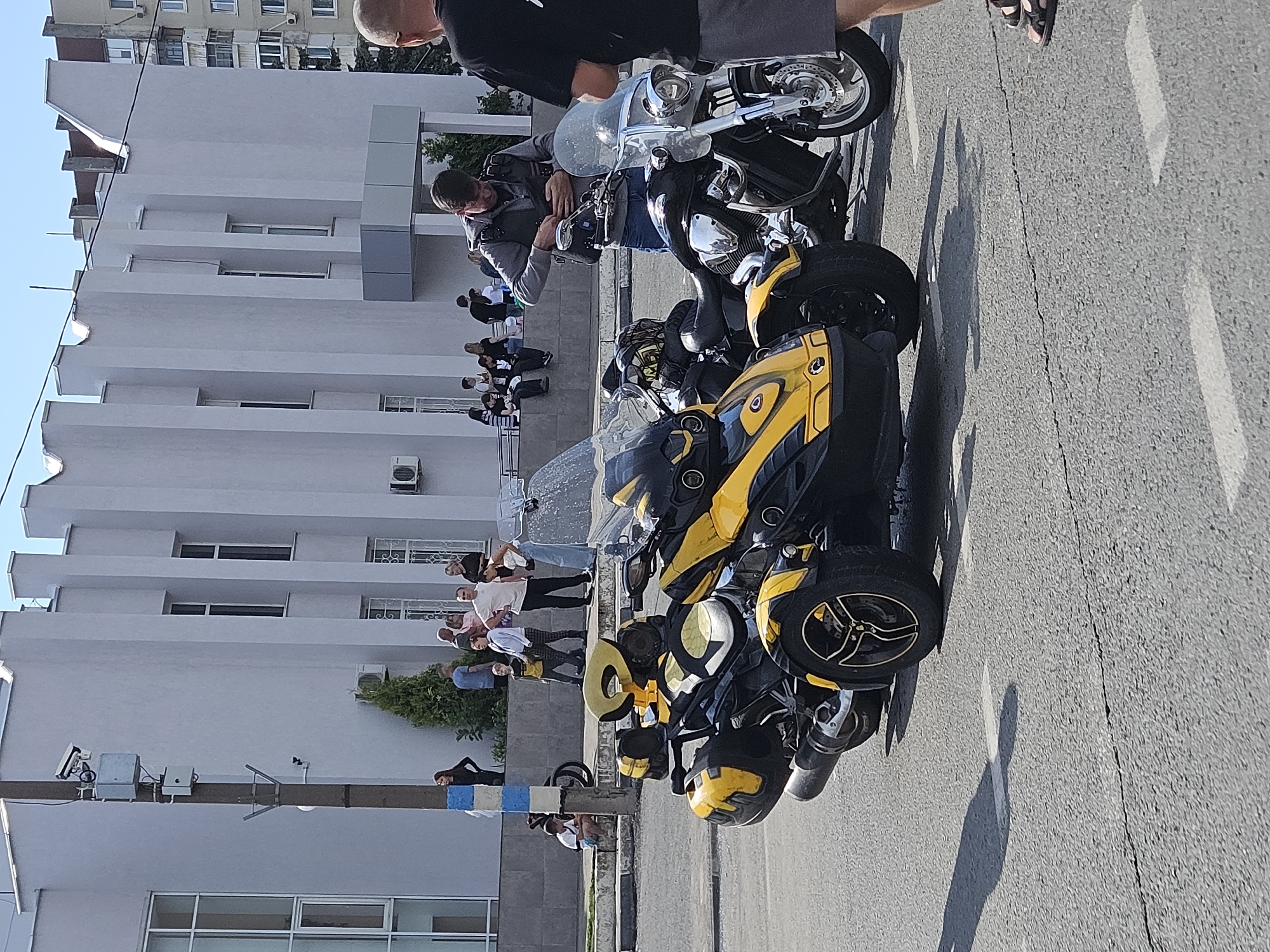 A medium shot shows a sunny daytime scene on an asphalt road in front of a light-colored building with a modern architectural style. In the foreground, a yellow and black three-wheeled motorcycle is parked, with its front angled slightly to the left. Behind it, a black motorcycle is also parked. A man in a gray vest over a gray hoodie and blue jeans is standing next to the black motorcycle, adjusting the zipper on his vest. To the right of the black motorcycle, the lower half of a person wearing a black t-shirt and gray shorts is visible, with their feet in sandals on the road. In the middle ground, a group of people are standing and sitting on a low concrete wall in front of the building. Some are looking at the motorcycles, while others are interacting amongst themselves. The building has multiple windows and vertical decorative elements on its facade. A wooden pole with electrical equipment and surveillance cameras is positioned to the left of the motorcycles. The overall scene appears to be an outdoor gathering or event.