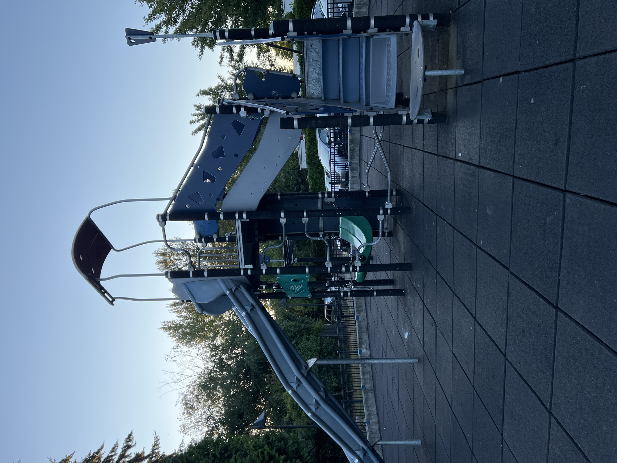 The image depicts a playground structure at dusk, with a clear blue sky overhead. The playground features a prominent blue and gray slide, stairs, and green swings. The ground is covered with black rubber tiles, and metal posts support the playground equipment. In the background, trees and parked cars are visible, suggesting an urban or suburban setting. The scene is peaceful and devoid of any people. Based on visual cues and common playground designs, this playground is likely located in a park or community area within the city of Kent, United States of America.