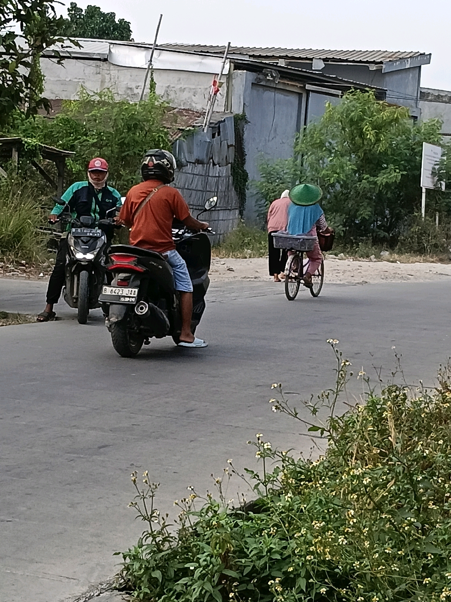 In the city of Belimbing, Indonesia, a man wearing a red cap and a green and black jacket rides a black motorcycle with a license plate reading "B 6423 JAA". He is followed by another person on a similar black motorcycle, wearing an orange shirt and blue shorts. To the right, two individuals are on a bicycle; one wearing a pink shirt and a blue head covering, and another person in pink pants and a green conical hat. They are riding past a building with a grey wall and a corrugated roof, partially obscured by green foliage. The scene takes place on a paved road.