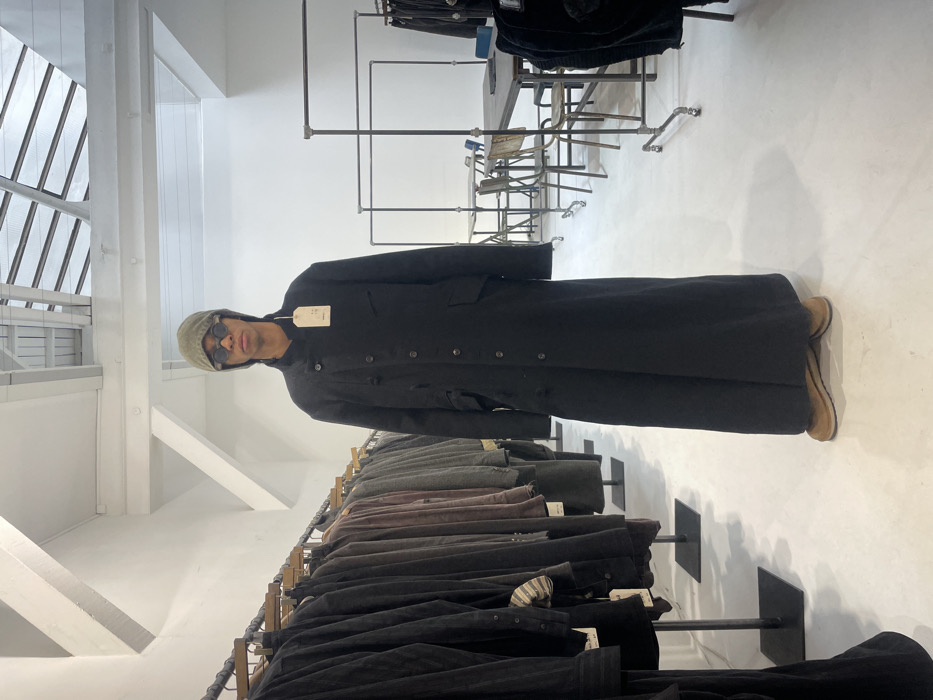The image portrays the interior of a fashion boutique or showroom situated in Paris, France. While a precise street address is not visually ascertainable, the architectural style—characterized by high white ceilings with exposed beams and rectangular skylights, clean white walls, and industrial-chic fixtures—is common in modern commercial spaces within fashion districts of the city.
A single adult male stands centrally, facing the camera. He is of slender build and wears a full-length, dark-colored (likely black) coat or robe extending to his ankles, featuring multiple visible buttons and pockets. A light-colored product tag hangs from the coat near his left shoulder. He wears a light olive-green or khaki beanie and small, round, dark-lensed sunglasses; his mouth is slightly open. His footwear consists of tan or light brown flat-soled shoes.
To the left, a linear arrangement of dark industrial-style clothing racks runs along a white wall, laden with numerous dark-toned garments (primarily black, dark grey, and muted earth tones), some bearing product tags. These racks stand on dark, square base plates on the floor. In the mid-ground to the right, several rectangular tables with dark tops and metal frames are visible, accompanied by various chairs, some appearing to be vintage industrial style with metal frames and wooden elements. Further right, additional lighter-colored industrial clothing racks also display dark garments.
The floor throughout the space is a light-colored, uniform polished surface (white or very light grey). The high ceiling, with its exposed beams and multiple skylights, provides ample natural light, contributing to a bright, minimalist, and industrial aesthetic. No active interactions or specific actions beyond posing are evident.