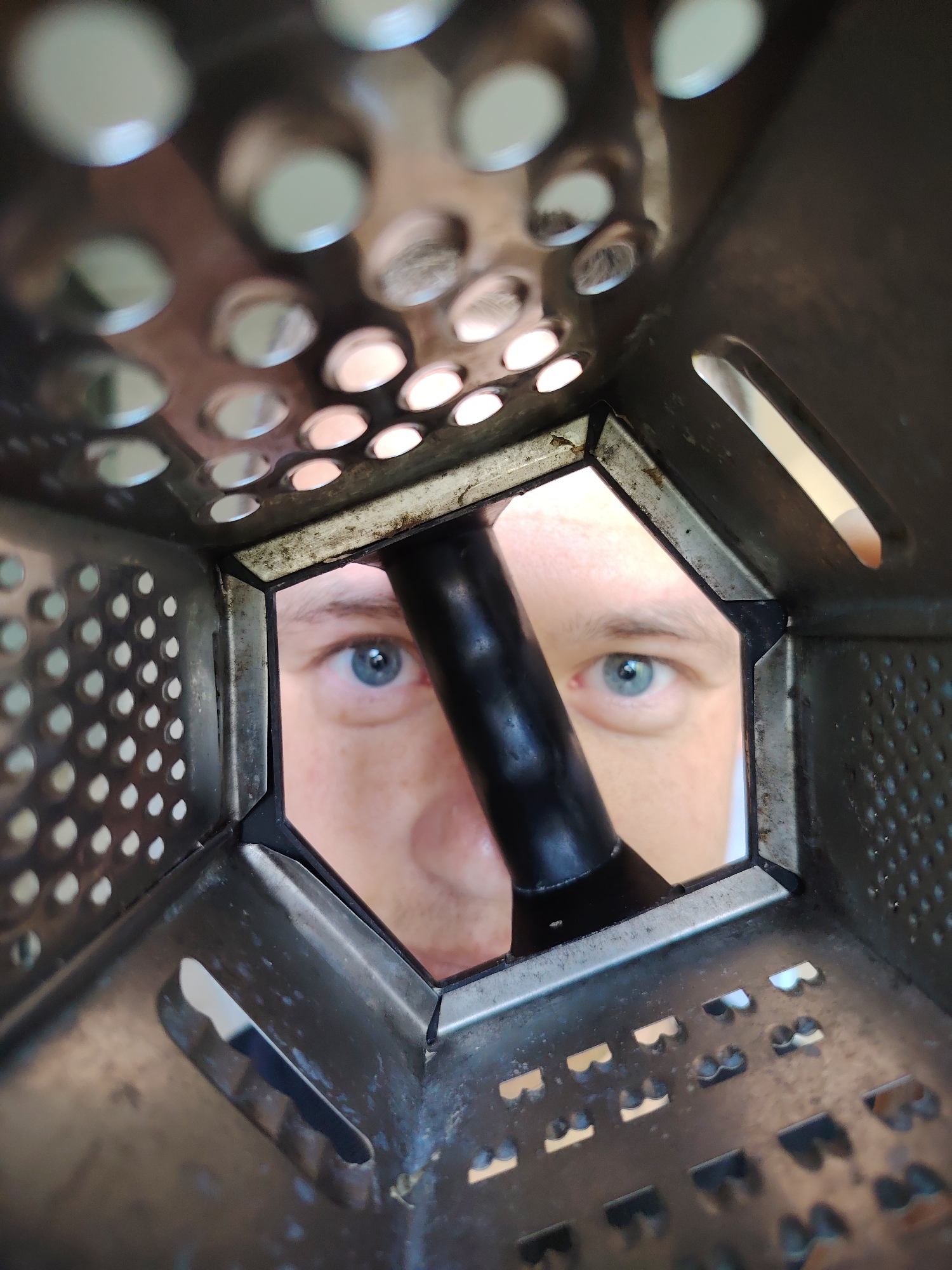 A close-up, slightly angled shot from inside a metal box grater reveals a person's face. The grater obscures much of the face, but two light blue eyes are visible, as well as the bridge of a nose and part of the face below. A black object, possibly a handle, bisects the visible facial area. The grater itself is metal, with various grating surfaces visible, including holes and slots. The background is indistinct. There is no indication of the location being Chaikovsky, Russia, in the image.