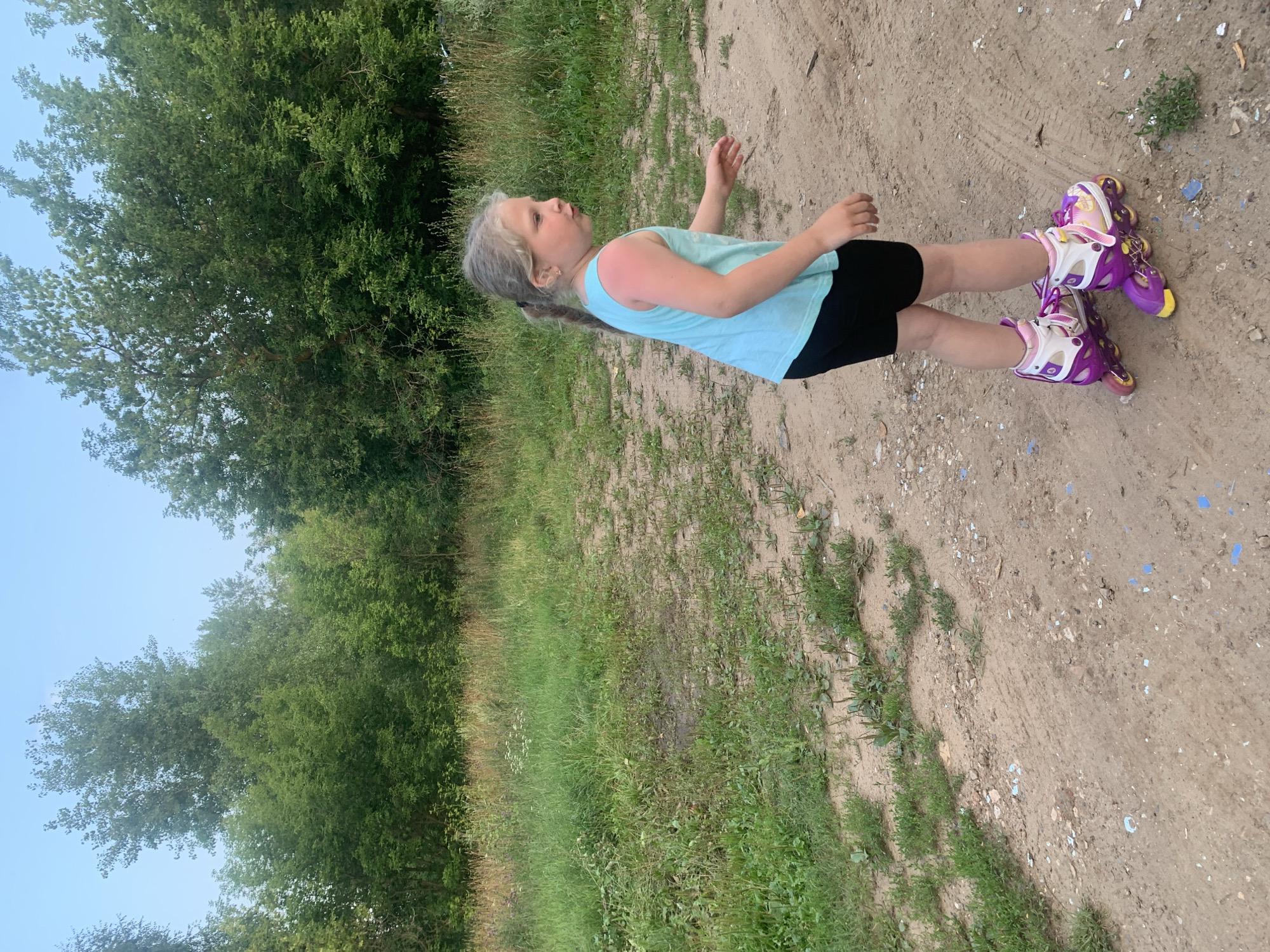 A young girl with blonde hair pulled back into a ponytail stands on a sandy path. She is wearing a light blue tank top, black shorts, and purple roller skates. Her right arm is raised slightly, and her left hand is extended forward. Her skin on her right arm appears sunburned. The girl is looking to her left with an open mouth, as if she is speaking or surprised.
The setting is outdoors, with lush green trees and grass bordering the path. The sky is a light blue, suggesting it is daytime, likely late afternoon or early evening as indicated by the presence of shadows. The ground is a mixture of sand and dirt, with small pebbles and some patches of green vegetation growing through. The overall impression is a casual, outdoor activity. The presence of the trees and the type of vegetation suggest a rural or park-like setting. There is no visible text in the image.