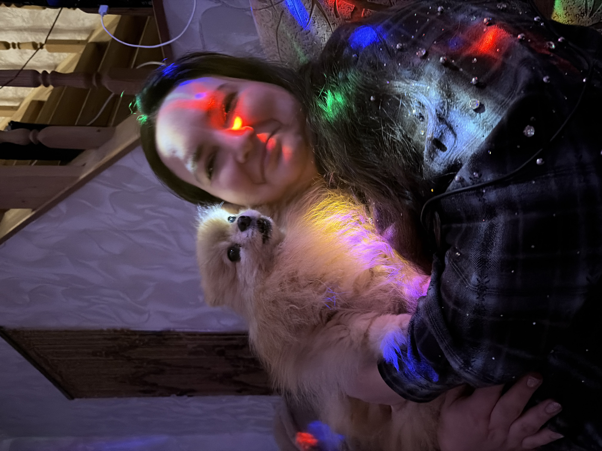 A woman is holding a small, fluffy dog, likely indoors. The scene appears to be illuminated by Christmas lights, casting a variety of colors on the woman and the dog. The woman is wearing a dark plaid shirt with small embellishments. A staircase is visible in the background. While the context suggests a domestic environment, there is no information to definitively identify this location as Pirogovo, Russia. Therefore, I cannot include the location in my description.