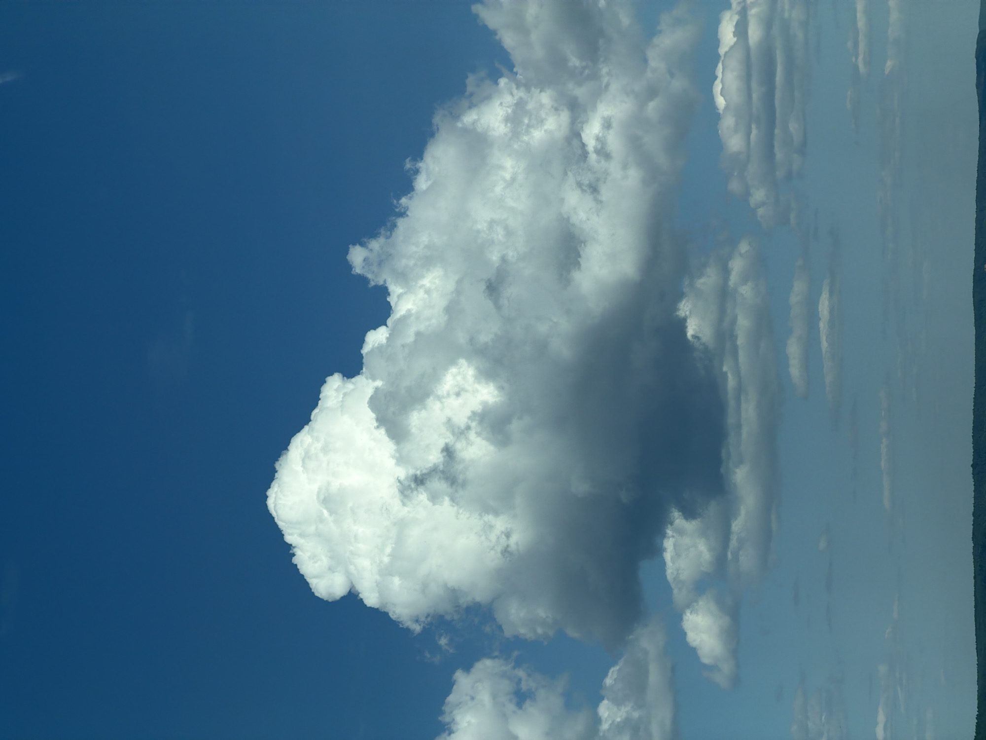 This image, captured in Chaikovsky, Russia, depicts a expansive sky dominated by cloud formations over a distant horizon. The upper two-thirds of the frame features a deep, clear blue sky. A large, prominent cumulus cloud formation occupies the central and left-mid section, exhibiting bright white, illuminated tops and darker, grey-shaded undersides, indicating significant depth and light interaction. To its right, other clouds, appearing as a mixture of cumulus and stratocumulus, extend horizontally, also showing variations in light and shadow. Towards the lower portion of the frame, near the horizon, the sky transitions to a lighter, paler blue, with several smaller, flatter cloud layers dispersed throughout. The lowest visible element is a thin, dark, undulating line representing a distant landmass or forest line, against which the lighter sky and lower clouds contrast. No people, specific objects, or actions are discernible within the frame. The scene is illuminated by natural daylight.