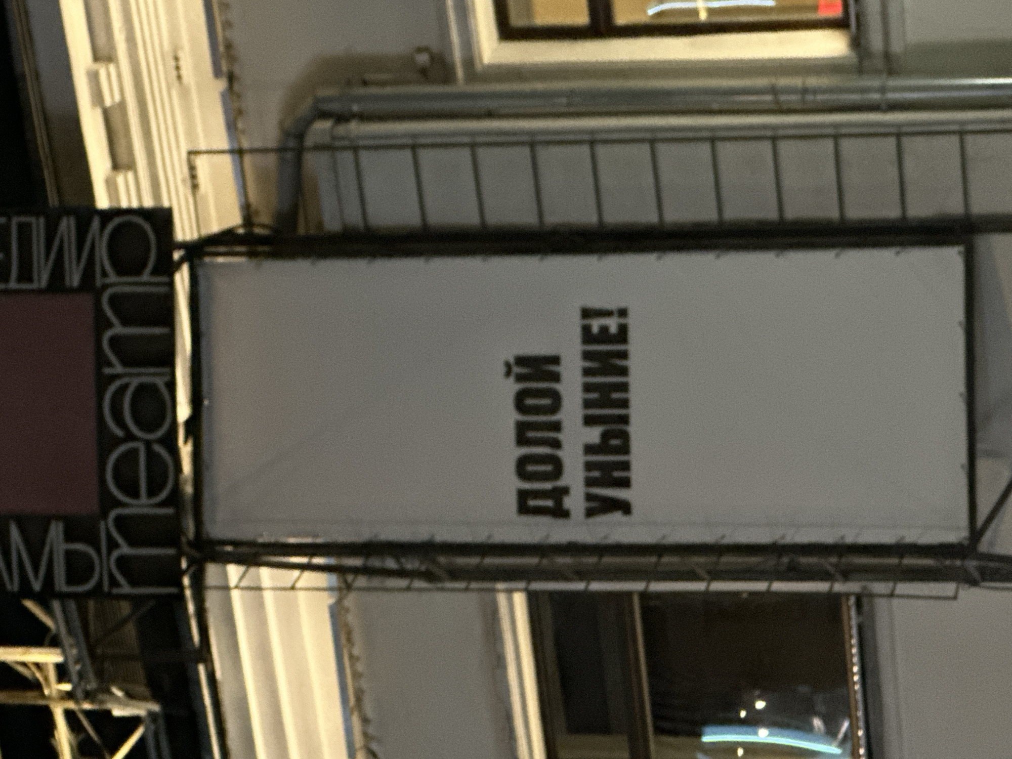 The image shows an outdoor sign in Moscow, Russia. The sign, which appears to be attached to a building, features the phrase "ДОЛОЙ УНЫНИЕ!" written vertically in black. The building has windows, some reflecting light. Part of another sign with Cyrillic text is also visible to the left.