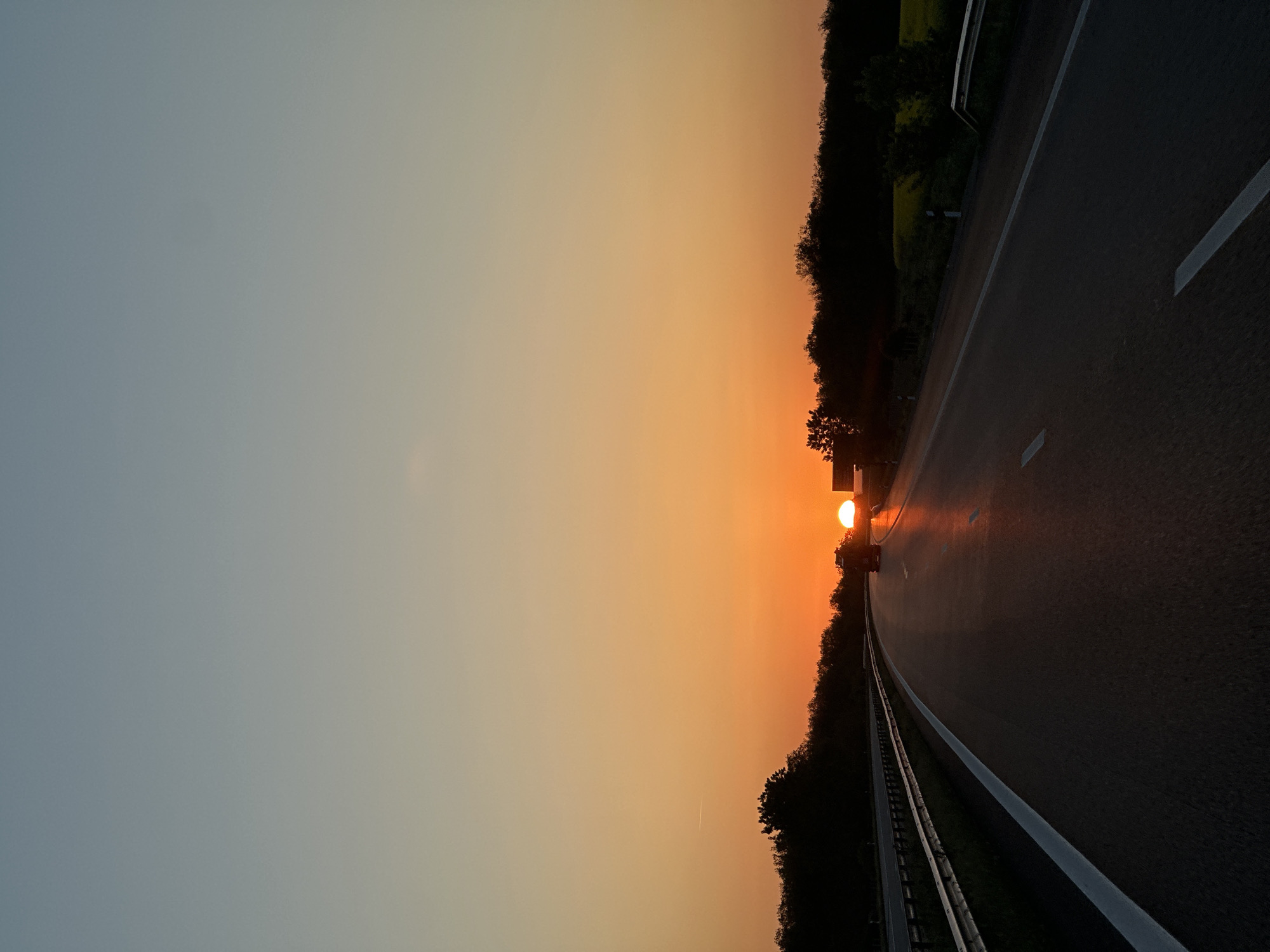 The image captures a highway scene at either sunset or sunrise, showcasing a dramatic sky with a gradient of colors. The sun, a bright orange orb, is positioned low on the horizon, emitting a powerful glare that washes over the center and right side of the image. The sky transitions from a vibrant orange and yellow near the sun to a calmer, light blue-grey higher up.
A dark asphalt road occupies the foreground and extends into the distance, with clear white lane markings and a solid white line along the shoulder. Guardrails are visible along the edge of the road, separating it from the surrounding landscape. Dense trees and foliage line both sides of the highway, appearing mostly in silhouette against the bright sky, with some glimpses of green further into the trees on the left. A small, dark shape, possibly a vehicle, is indistinctly visible on the road, directly in the path of the sun's intense glare. The overall atmosphere suggests a serene moment during travel. No text is visible in the image. The location is Боздуганово, Bulgaria.
